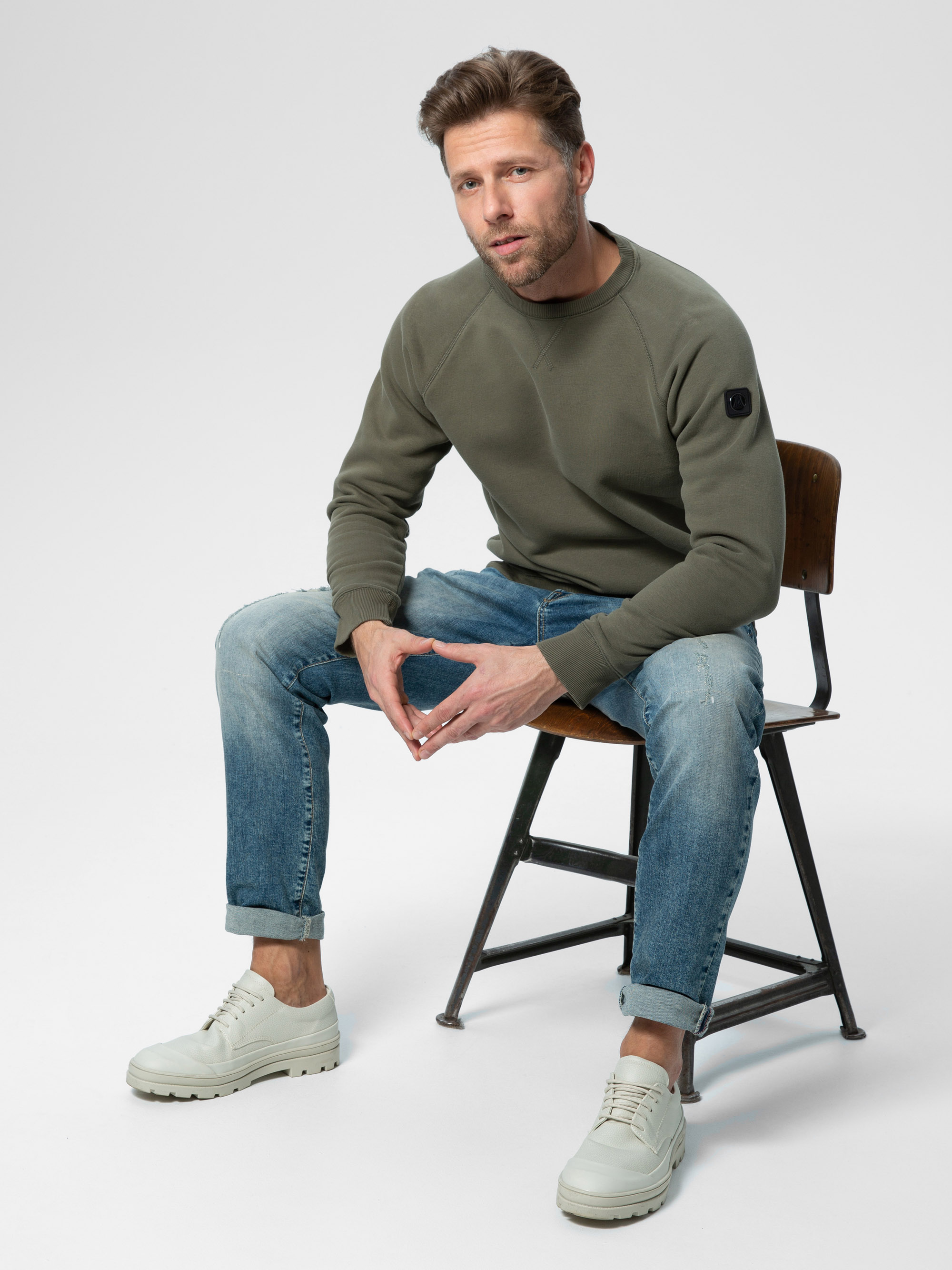 Ein Mann mit kurzen braunen Haaren und gestutztem Bart sitzt auf einem Holzstuhl und trägt ein grünes Sweatshirt, hellblaue Jeans und weiße, klobige Schuhe vor einem schlichten weißen Hintergrund.