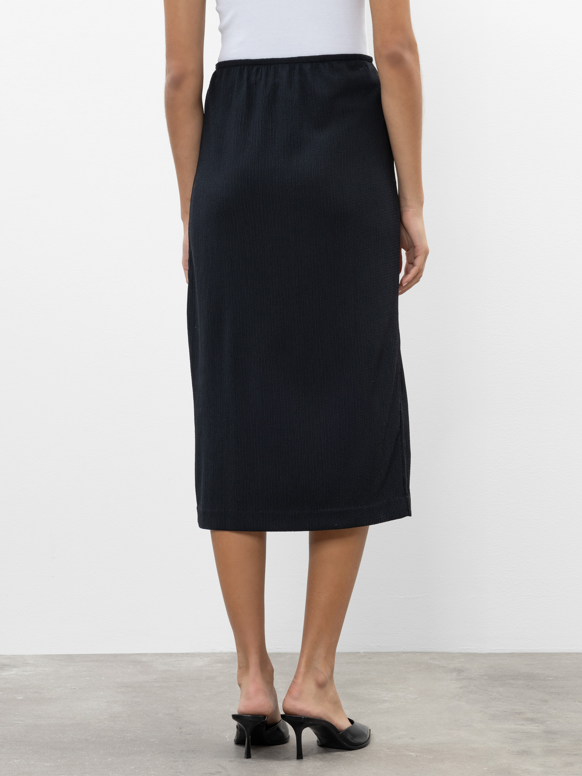 Eine Person, die ein weißes Oberteil, einen schwarzen Midirock und schwarze Pantoletten mit Absatz trägt, steht mit dem Gesicht nach unten auf einem Betonboden vor einer weißen Wand.