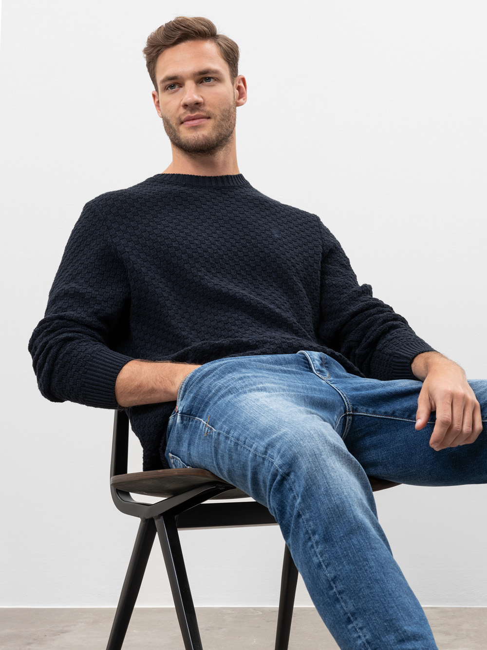 Ein Mann mit kurzen braunen Haaren, der einen dunklen strukturierten Pullover und blaue Jeans trägt, sitzt lässig auf einem schwarzen Stuhl vor einem schlichten weißen Hintergrund.