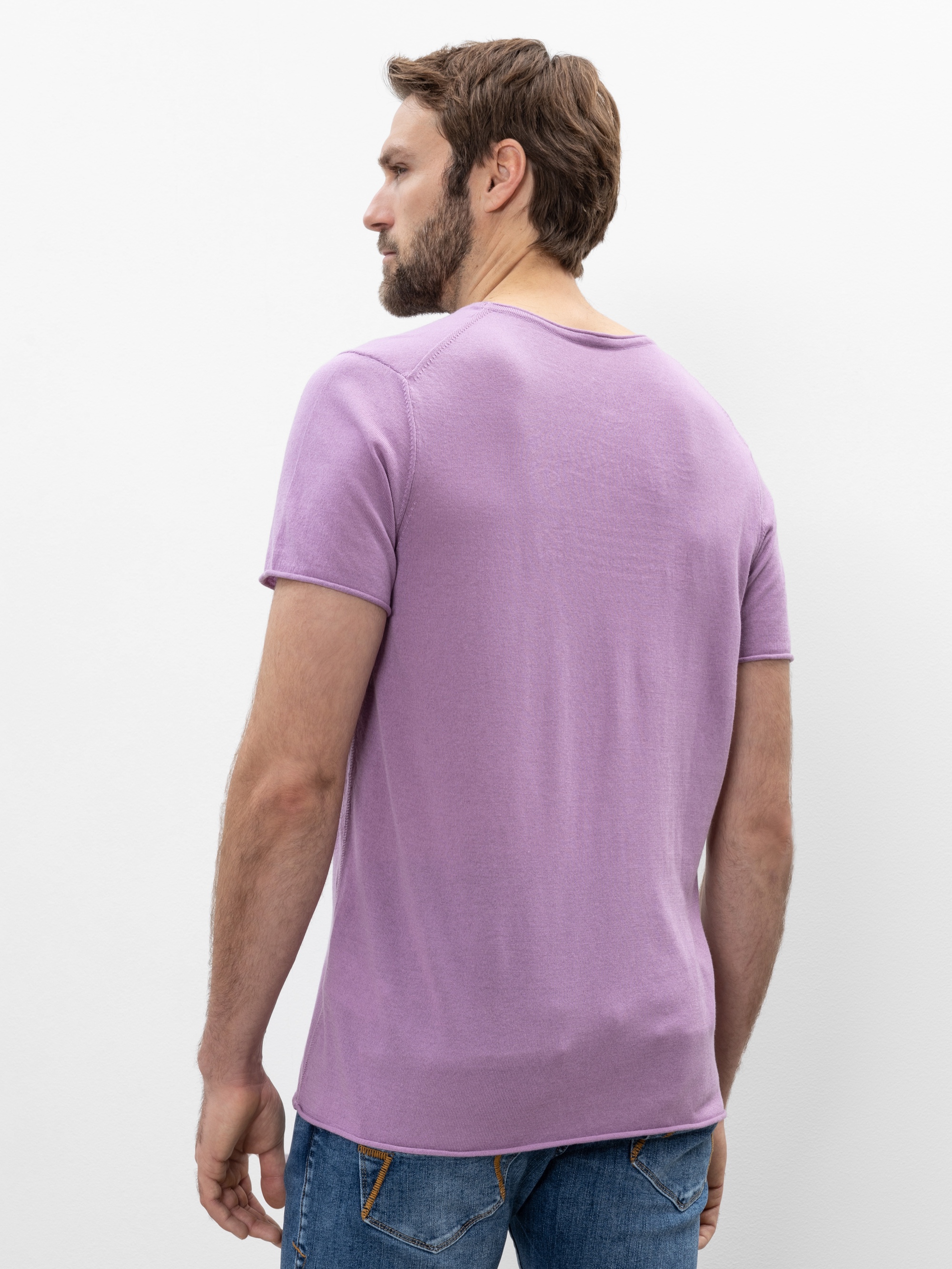 Ein Mann mit kurzen braunen Haaren und Bart, der ein lavendelfarbenes kurzärmeliges T-Shirt und blaue Jeans trägt, steht mit dem Gesicht zur Kamera vor einem schlichten weißen Hintergrund.