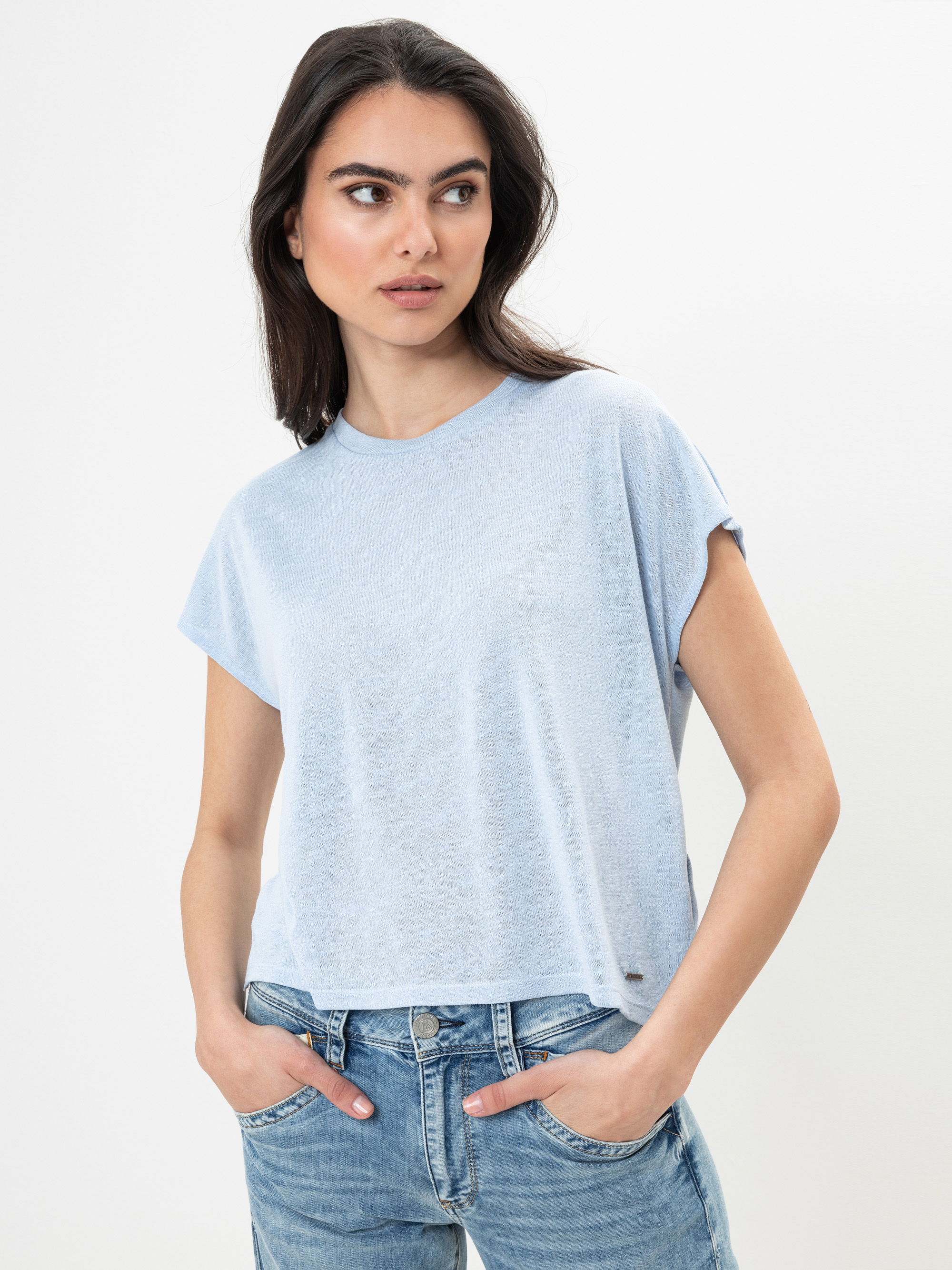 Eine Frau mit langen dunklen Haaren trägt ein hellblaues, kurzärmeliges, locker sitzendes T-Shirt und blaue Jeans. Sie hat die Hände in den Taschen und blickt nach links vor einem schlichten weißen Hintergrund.