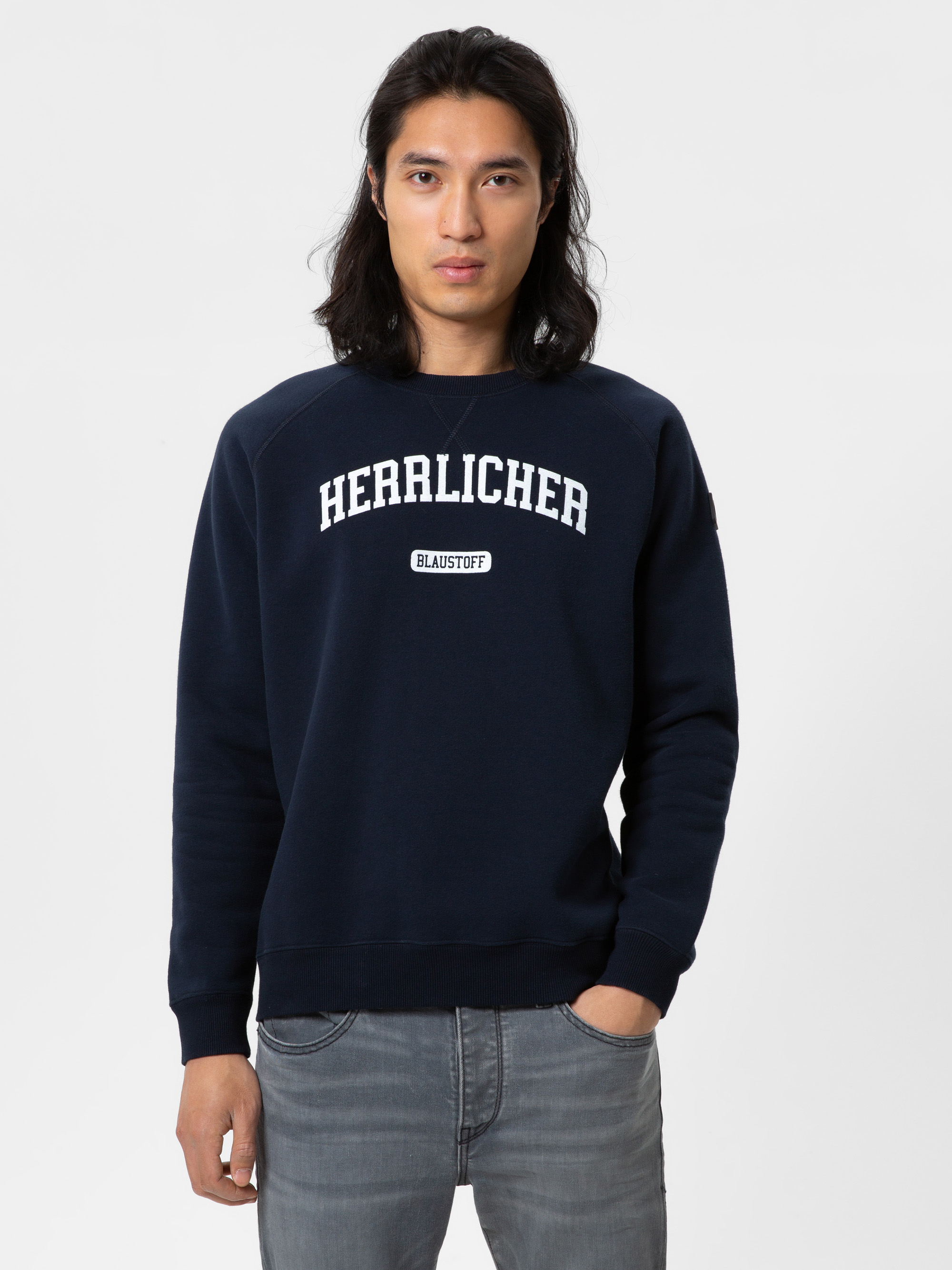 Eine Person mit langen dunklen Haaren trägt ein marineblaues Sweatshirt mit der Aufschrift HERRLICHER und BLAUSTOFF auf der Vorderseite, gepaart mit einer grauen Jeans, und steht vor einem schlichten weißen Hintergrund.
