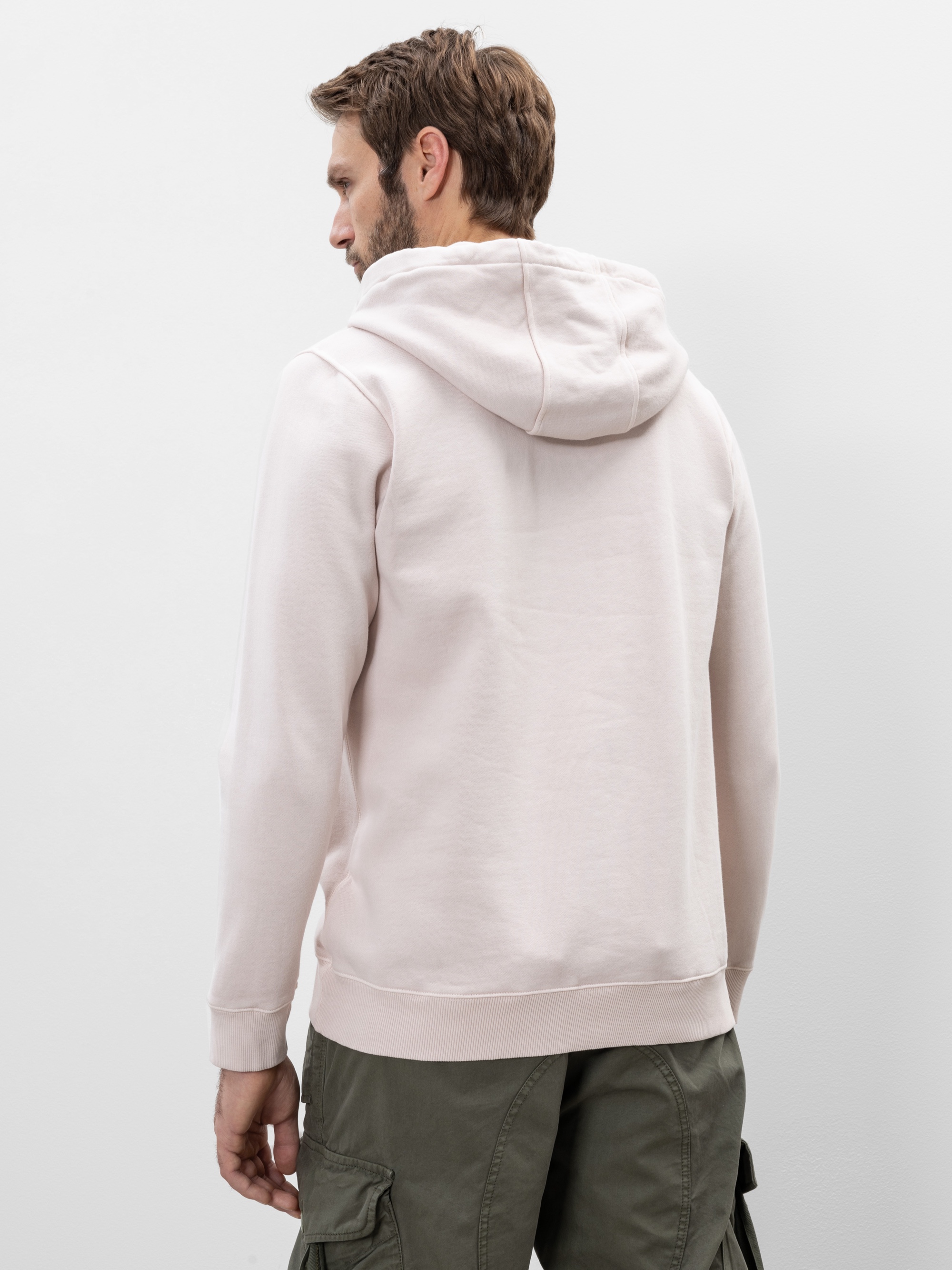 Ein Mann mit hellbraunem Haar steht mit dem Rücken zur Kamera, trägt ein blassrosa Kapuzensweatshirt und eine olivgrüne Cargohose vor einem schlichten hellen Hintergrund.
