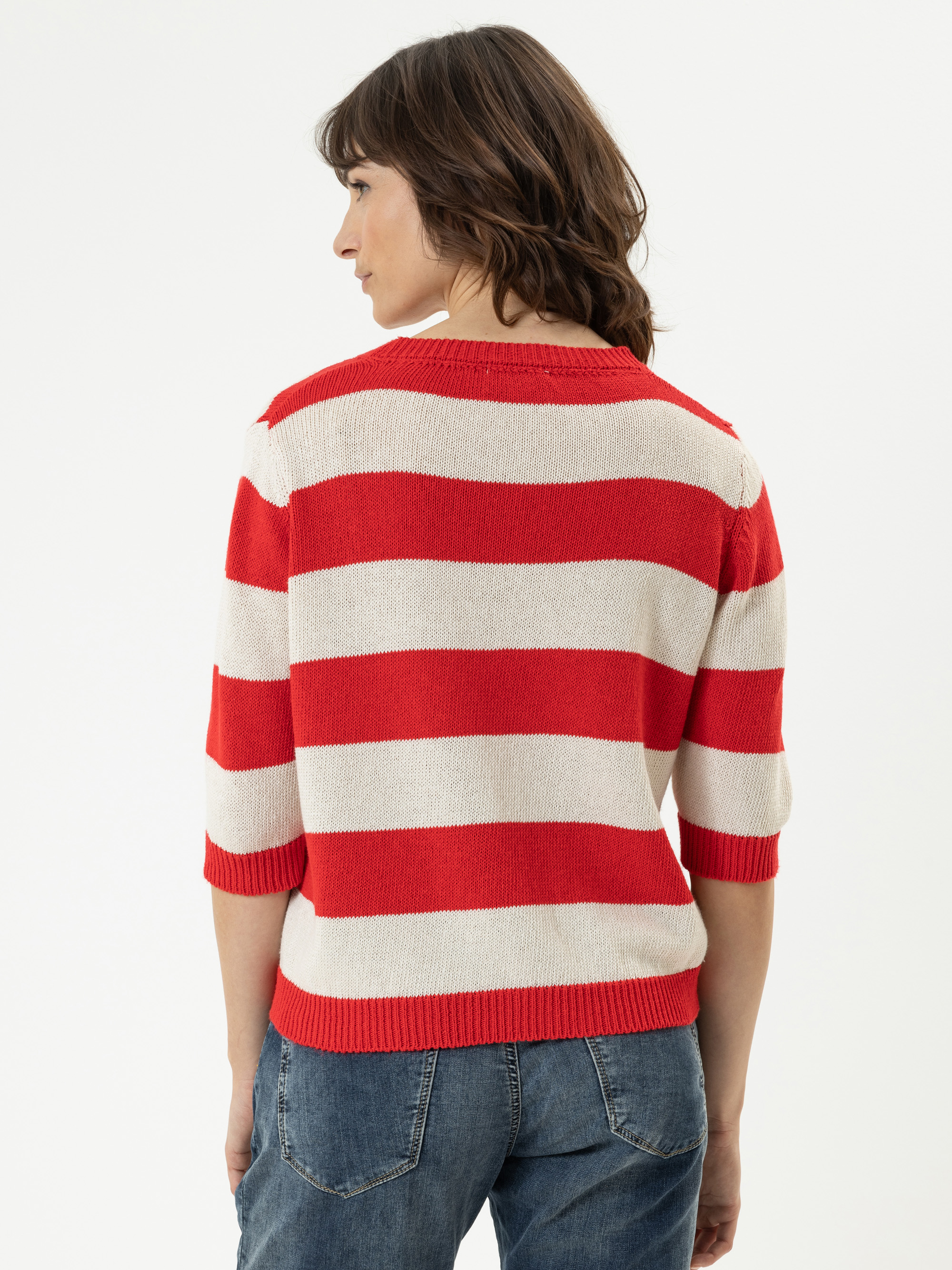 Eine Person mit schulterlangen braunen Haaren, die einen rot-weiß gestreiften kurzärmeligen Pullover und blaue Jeans trägt, steht mit dem Rücken zur Kamera vor einem schlichten weißen Hintergrund.