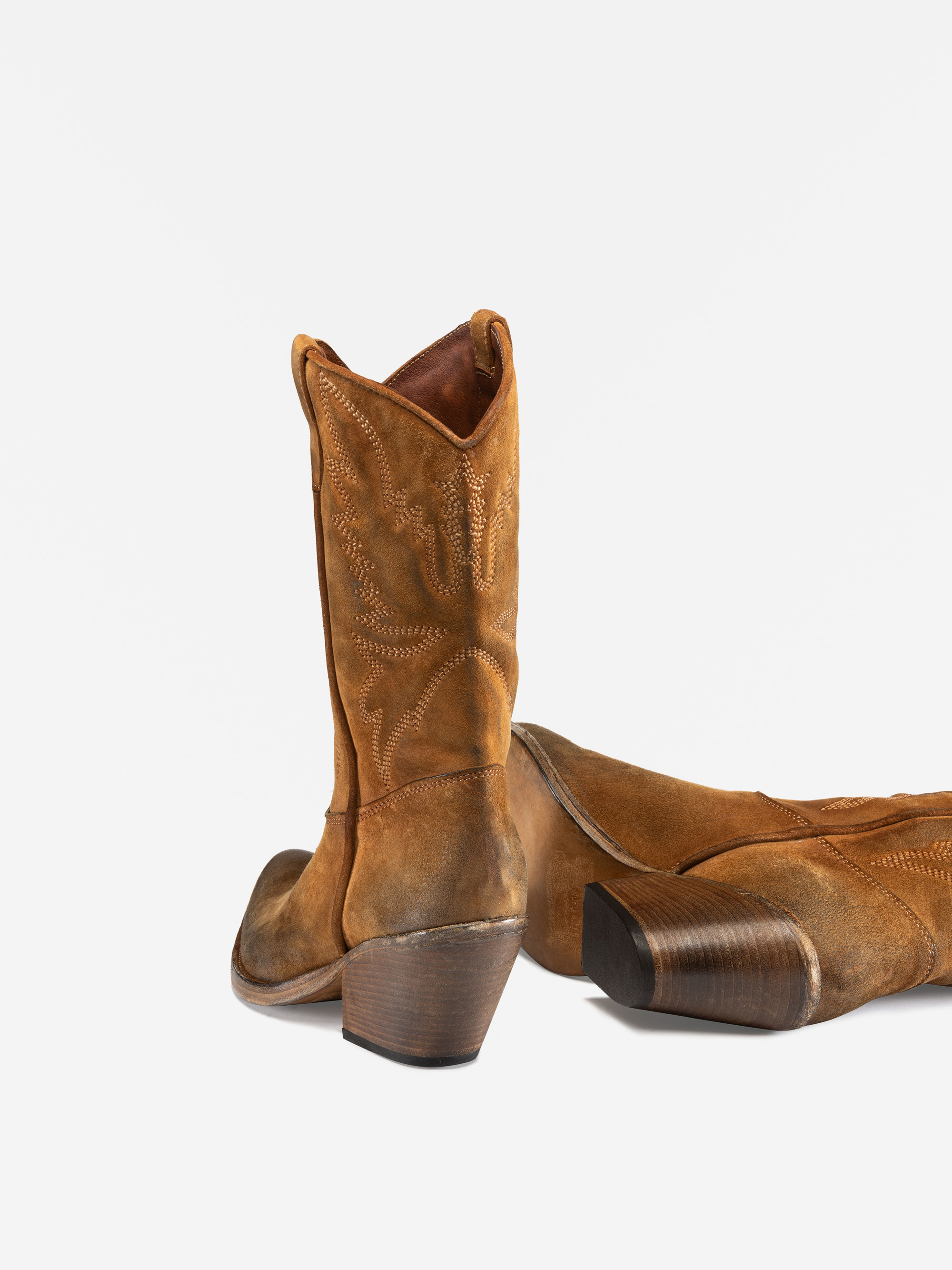 Ein Paar hohe, braune Cowboystiefel aus Wildleder mit Ziernähten und schrägen Holzabsätzen, wobei ein Stiefel aufrecht steht und der andere auf der Seite liegt, auf einem weißen Hintergrund.