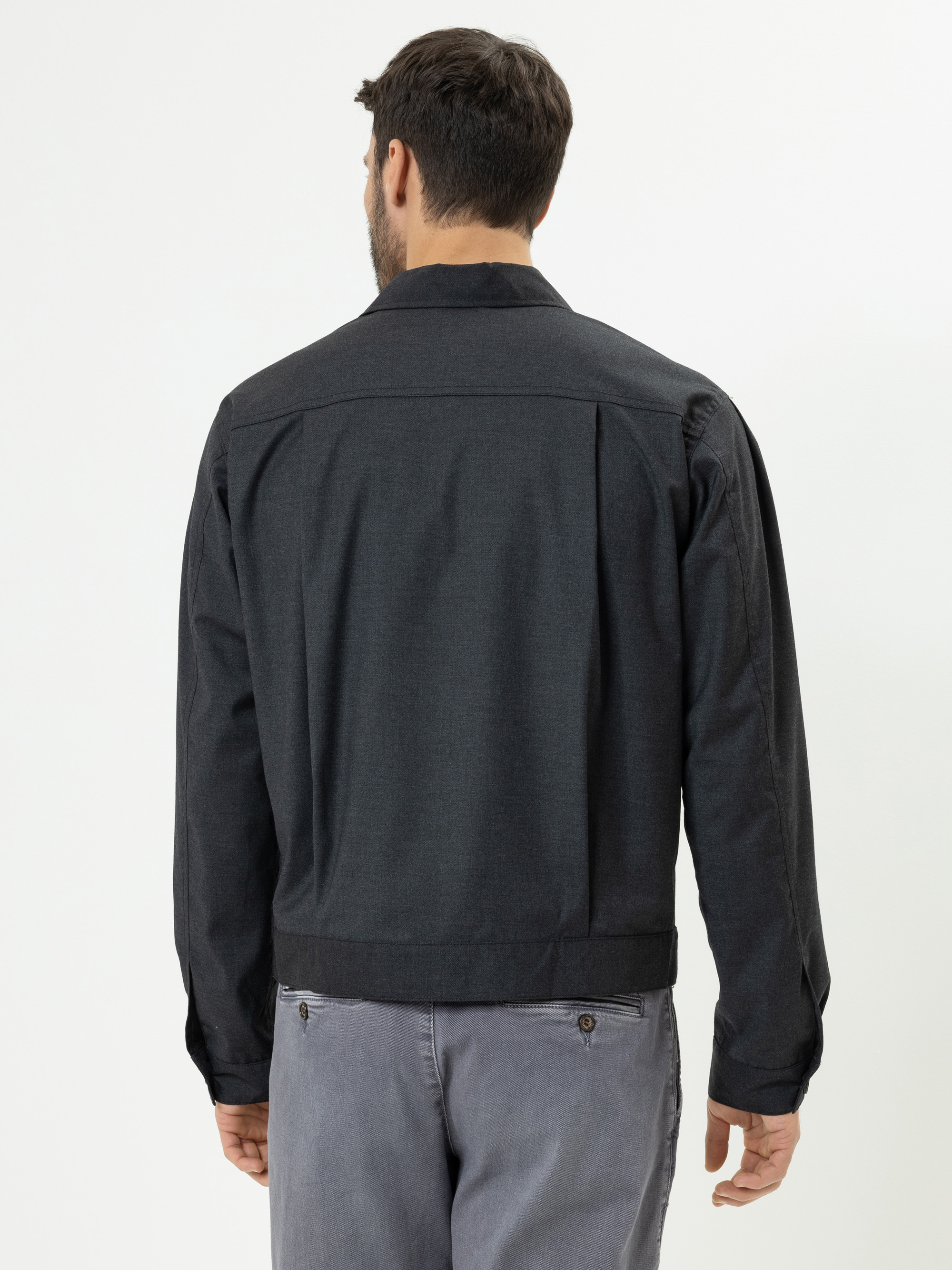 Ein Mann mit kurzen dunklen Haaren, von hinten gesehen, trägt eine dunkelgraue Jacke und hellgraue Jeans und steht vor einem schlichten weißen Hintergrund.