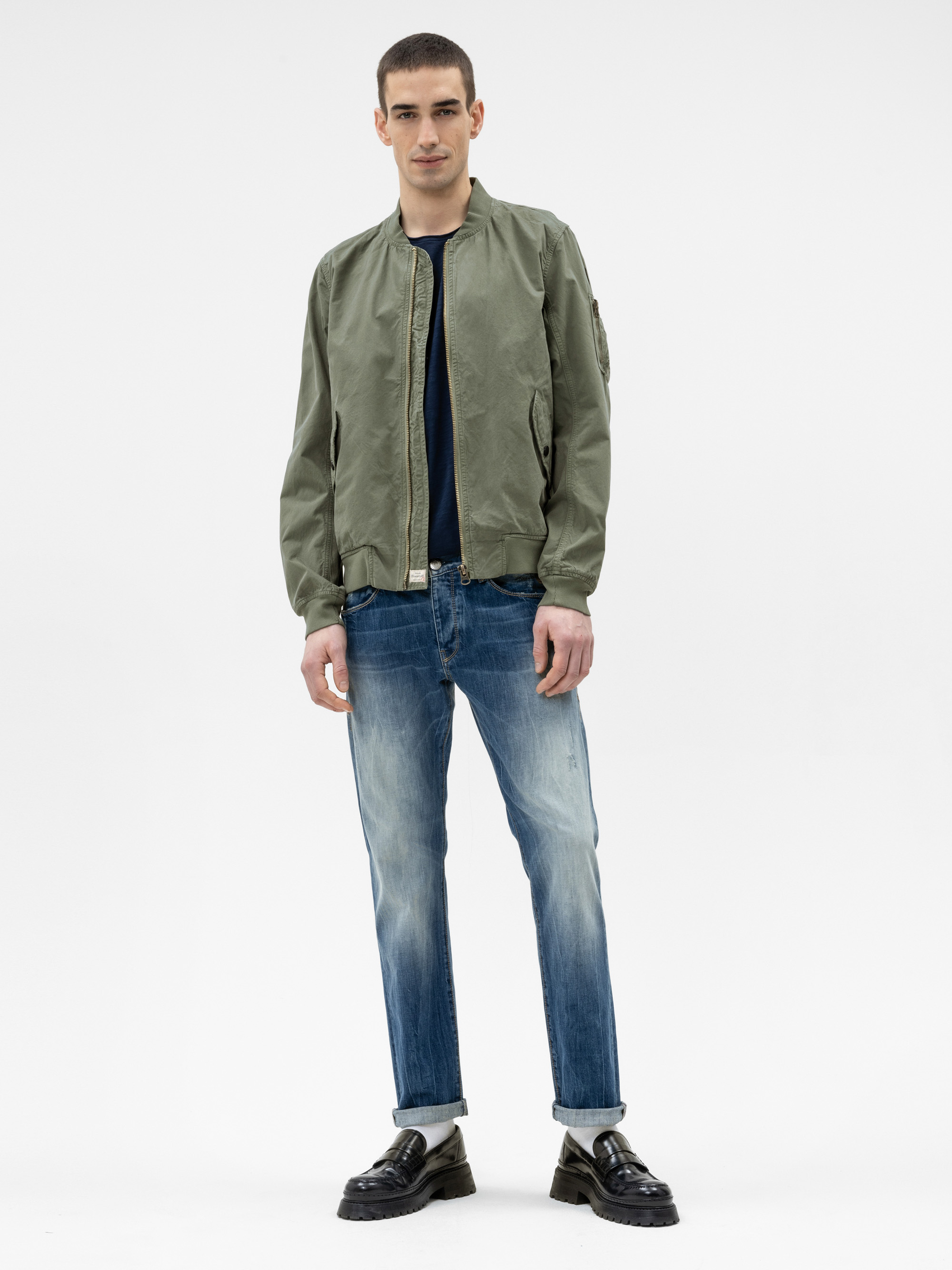 Ein junger Mann steht vor einem schlichten weißen Hintergrund und trägt eine hellolivfarbene Bomberjacke, ein dunkles T-Shirt, verblichene Bluejeans und schwarze Slipper. Er blickt nach vorne und hat die Hände an den Seiten.