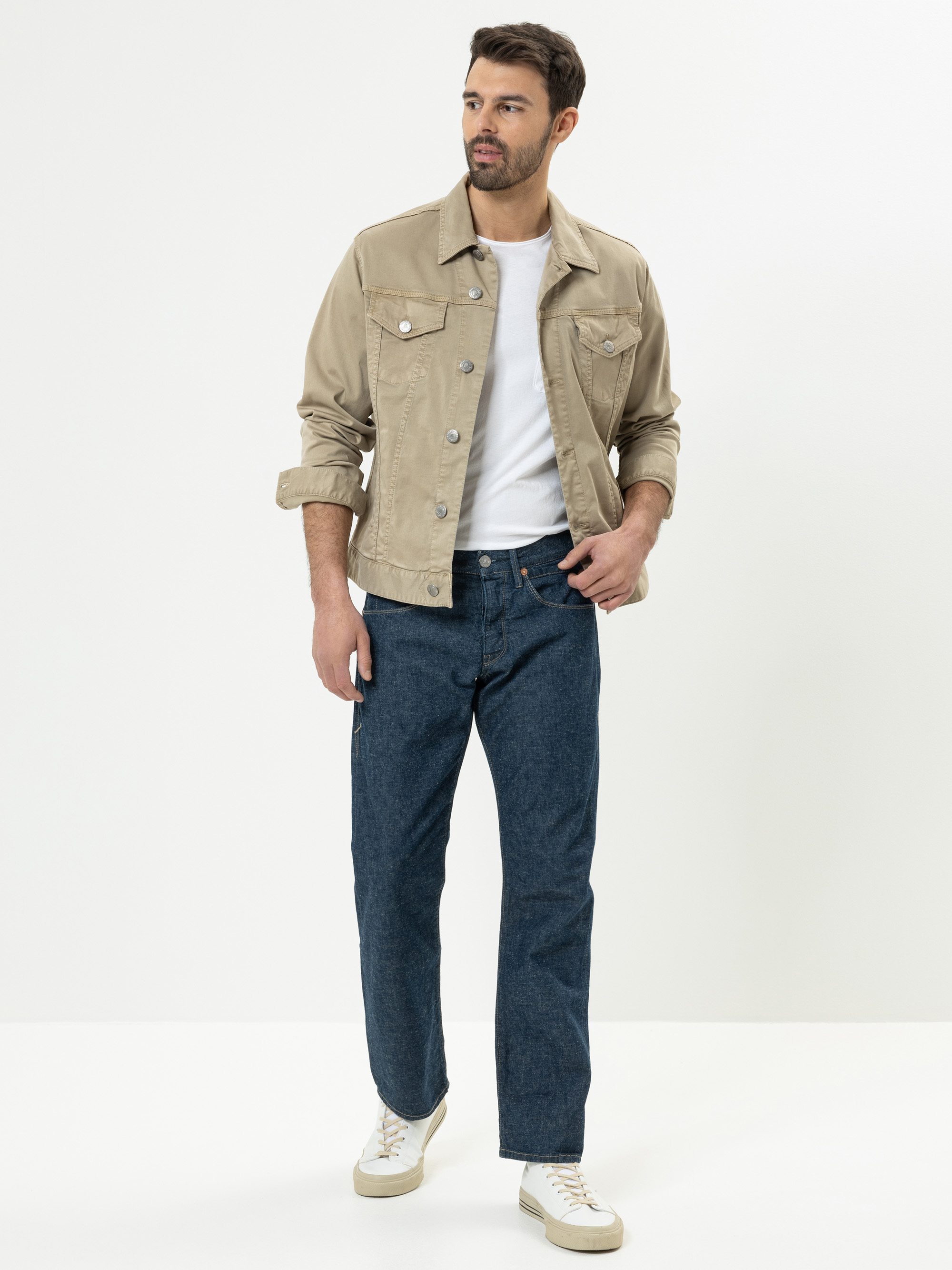 Ein Mann steht vor einem schlichten weißen Hintergrund und trägt eine beige Jeansjacke über einem weißen T-Shirt, blaue Jeans und weiße Turnschuhe. Er hat kurzes dunkles Haar und einen Bart und blickt leicht zur Seite.