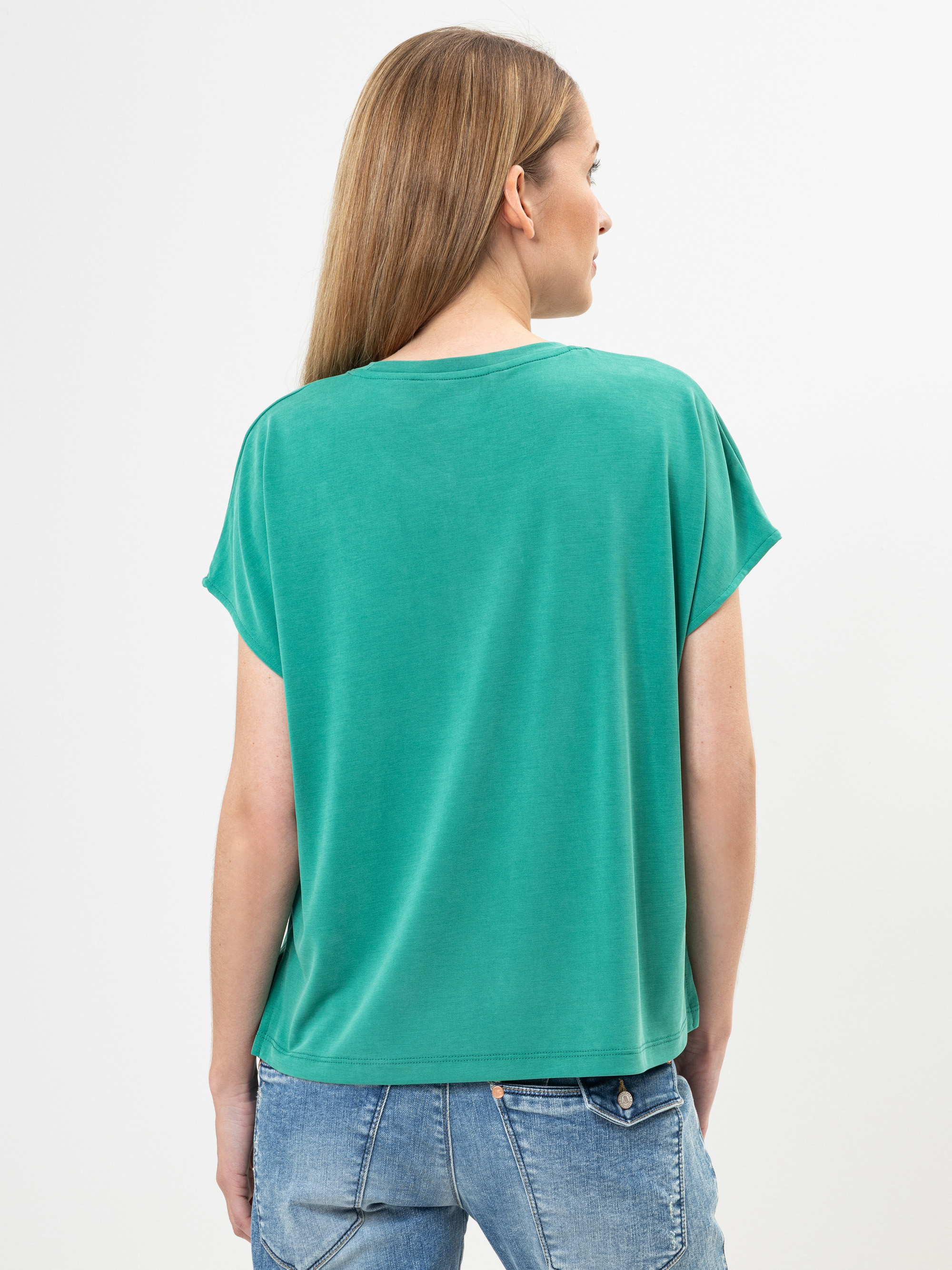 Eine Person mit langem, glattem, hellbraunem Haar steht mit dem Rücken zur Kamera und trägt ein locker sitzendes, grünes, kurzärmeliges Oberteil und hellblaue Denim-Jeans. Der Hintergrund ist einfarbig weiß.