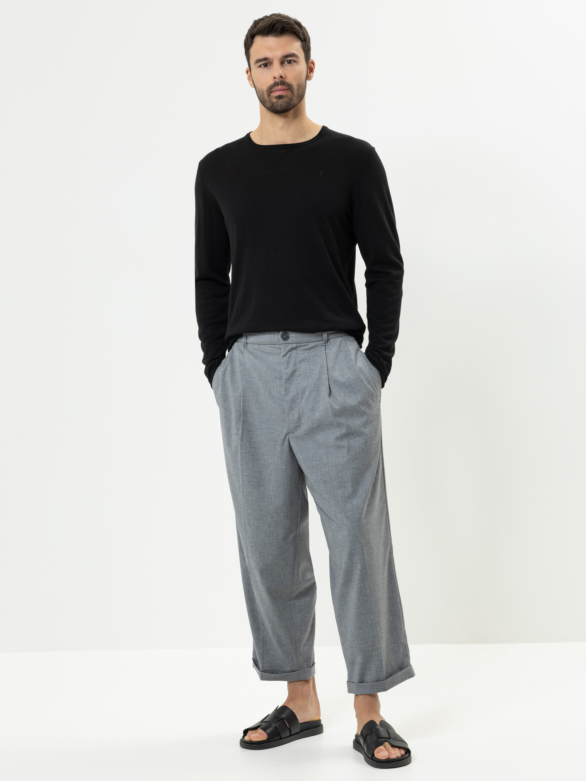 Ein Mann steht vor einem weißen Hintergrund und trägt ein schwarzes langärmeliges Hemd, eine weite graue Hose und schwarze Sandalen. Er hat die Hände in den Taschen und schaut mit neutralem Blick geradeaus.