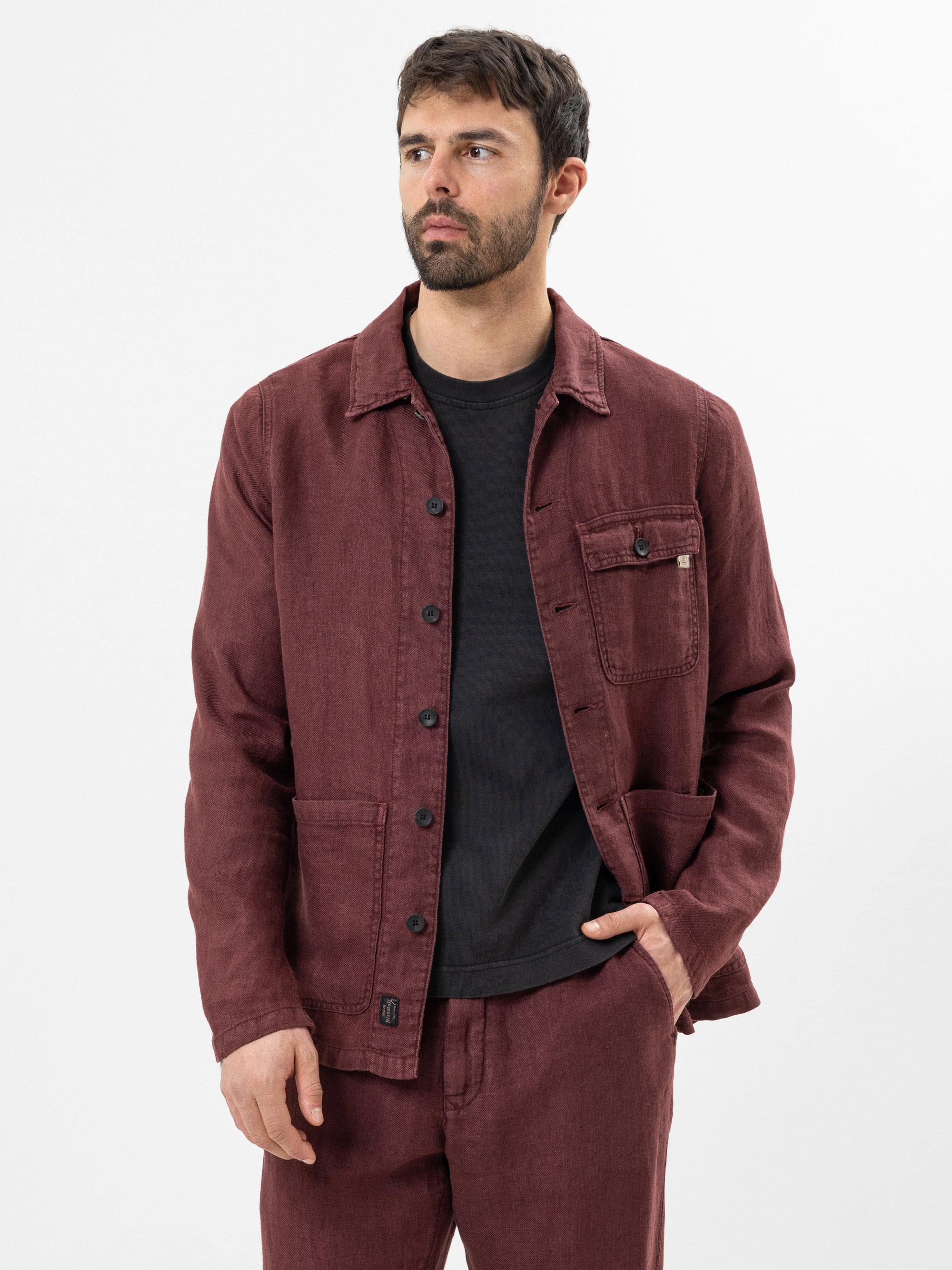 Ein Mann mit kurzen dunklen Haaren und einem Bart trägt eine kastanienbraune Button-up-Jacke über einem schwarzen T-Shirt und eine dazu passende kastanienbraune Hose. Er steht vor einem schlichten weißen Hintergrund und schaut leicht zur Seite.