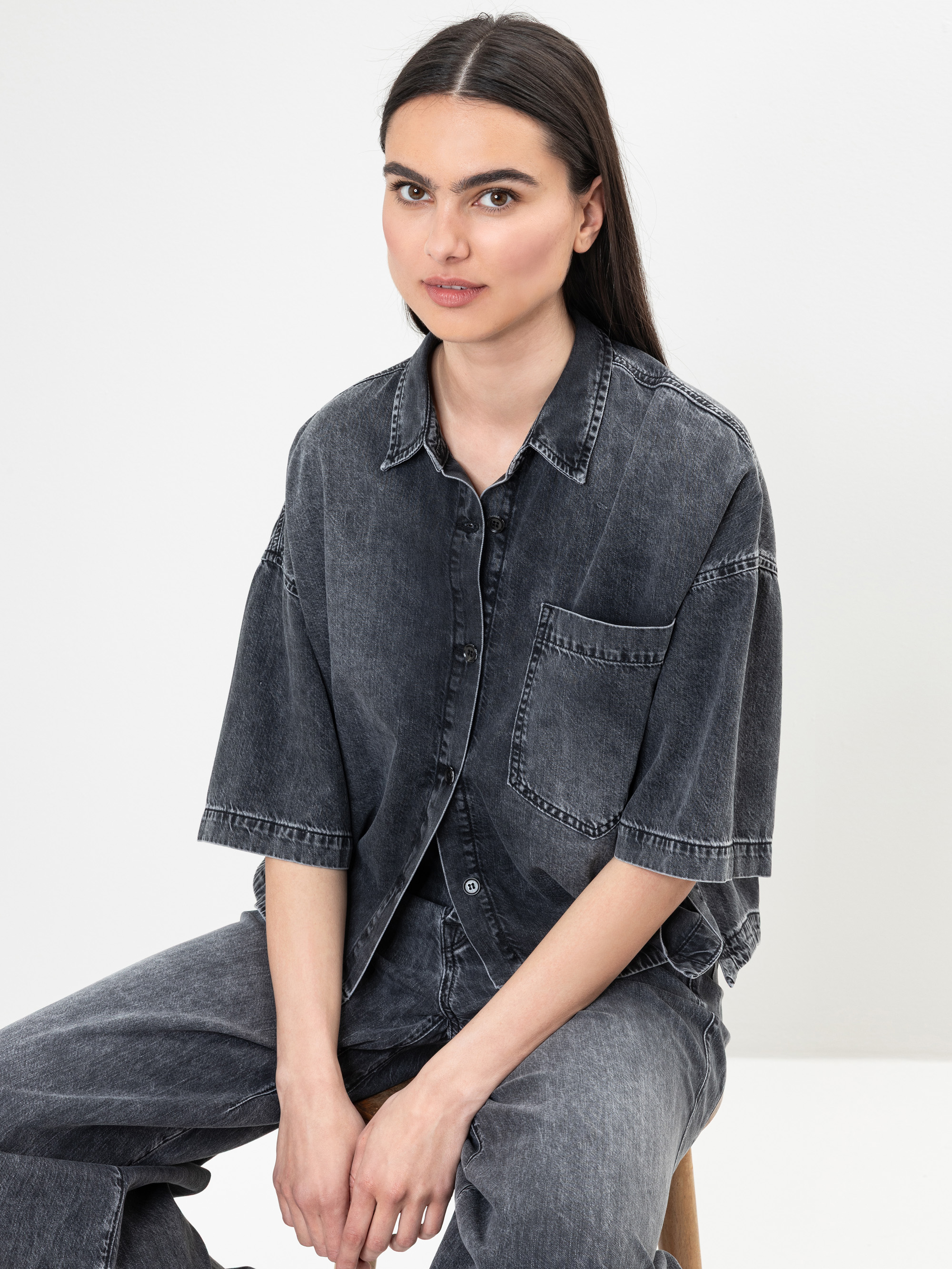 Eine Frau mit langen dunklen Haaren, die ein locker sitzendes, kurzärmeliges schwarzes Jeanshemd und eine passende Jeans trägt, sitzt auf einem Holzhocker vor einem schlichten weißen Hintergrund.