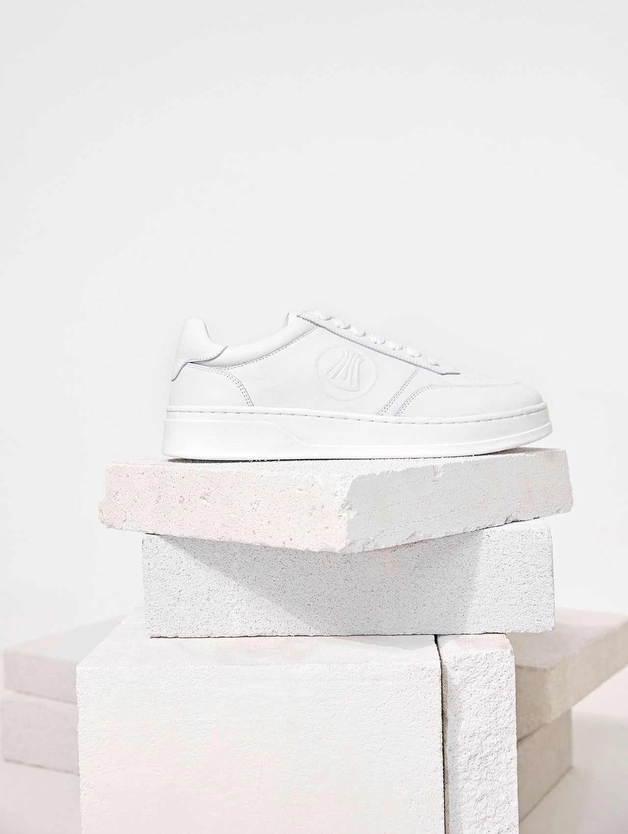 Ein weißer Turnschuh mit minimalem Design wird auf gestapelten weißen Betonblöcken vor einem schlichten weißen Hintergrund präsentiert.