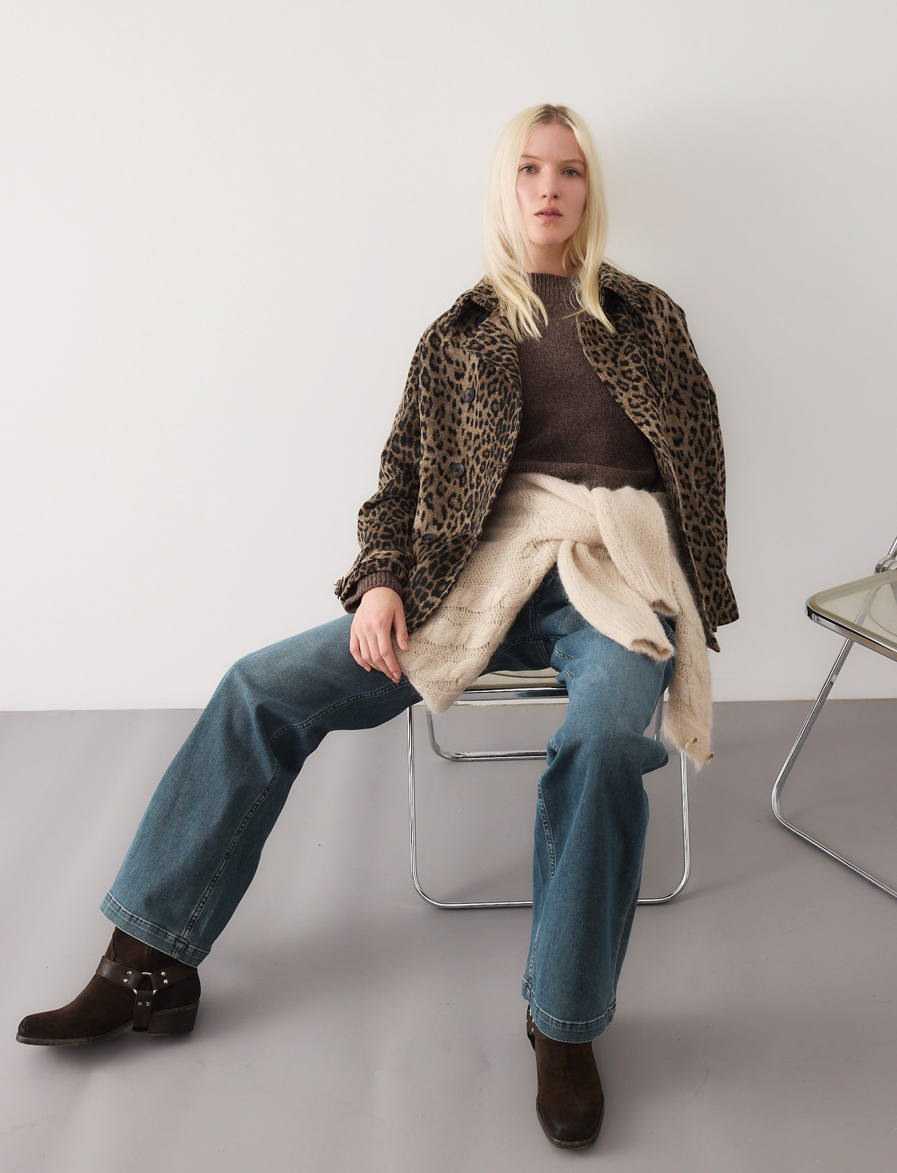 Eine Frau mit langen blonden Haaren sitzt auf einem hellen Stuhl und trägt eine Jacke mit Leopardenmuster, einen braunen Pullover, blaue Jeans und dunkle Stiefel. Ein beiger Pullover ist um ihre Taille gebunden. Ein weiterer leerer Stuhl steht neben ihr.