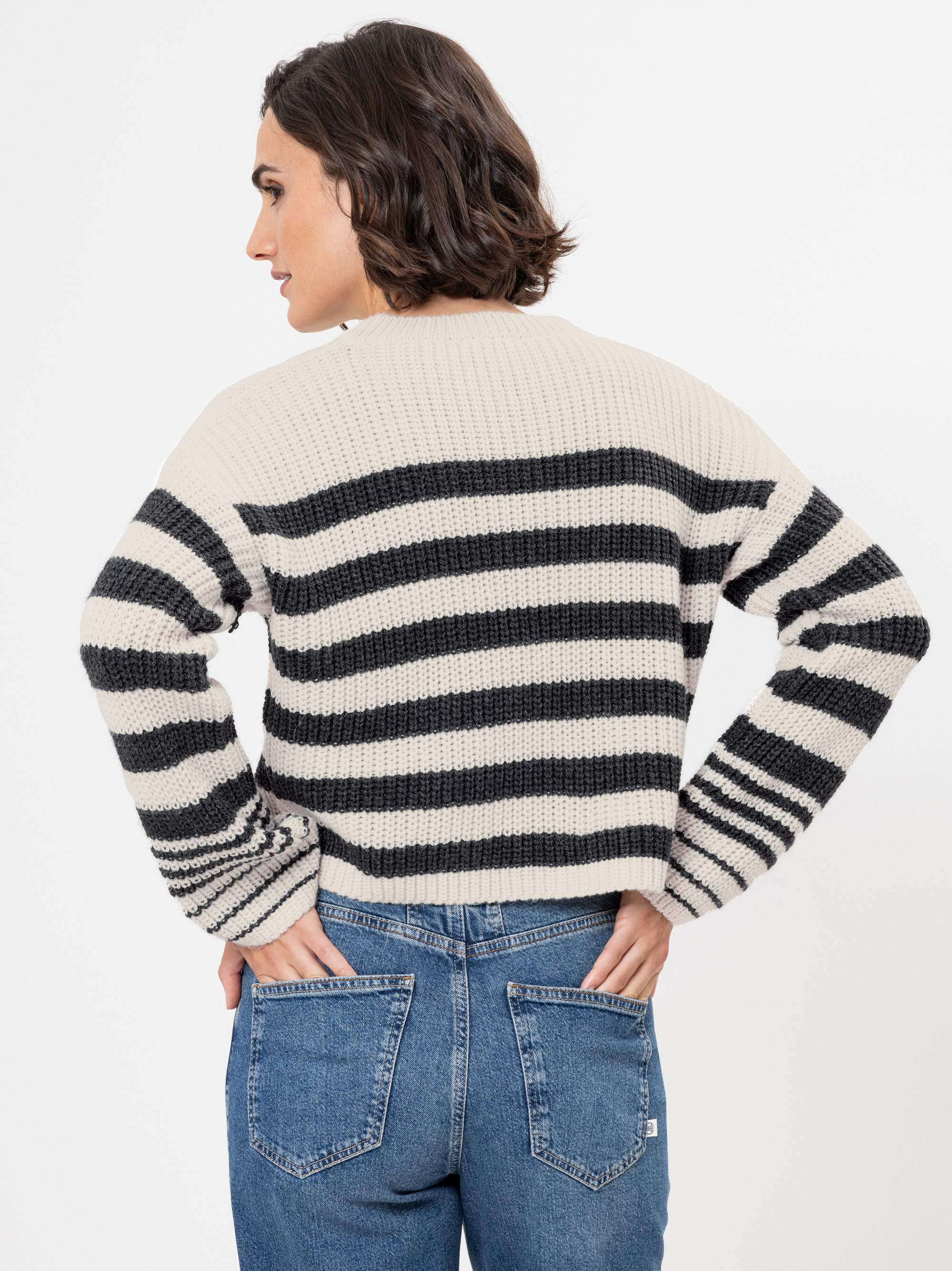 Eine Person mit kurzen braunen Haaren, die einen cremefarbenen und schwarz gestreiften Strickpullover und blaue Jeans trägt, steht mit dem Rücken zur Kamera und den Händen in den Hüften vor einem schlichten weißen Hintergrund.