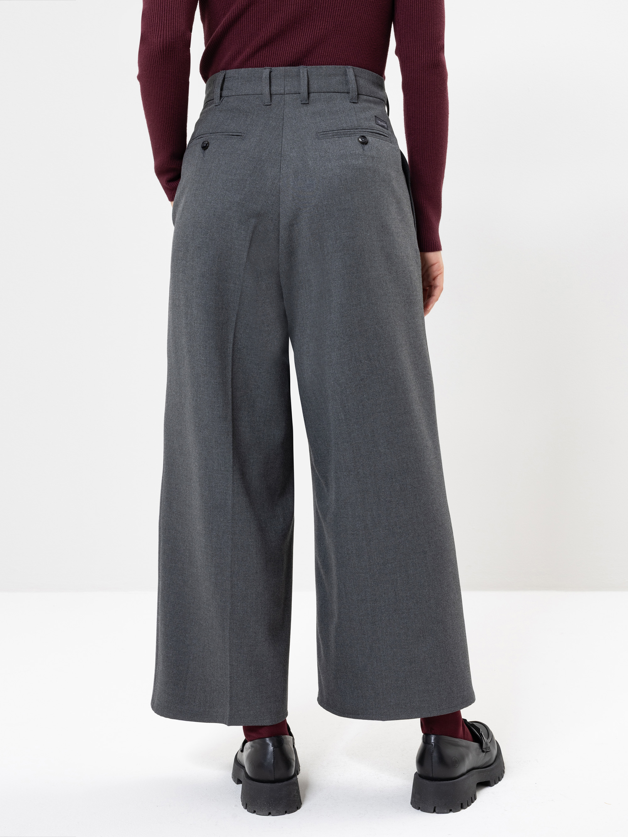 Eine Person, die eine graue Hose mit weitem Bein, ein weinrotes langärmeliges Oberteil, weinrote Socken und schwarze Schuhe trägt, wird von hinten vor einem schlichten weißen Hintergrund gezeigt.