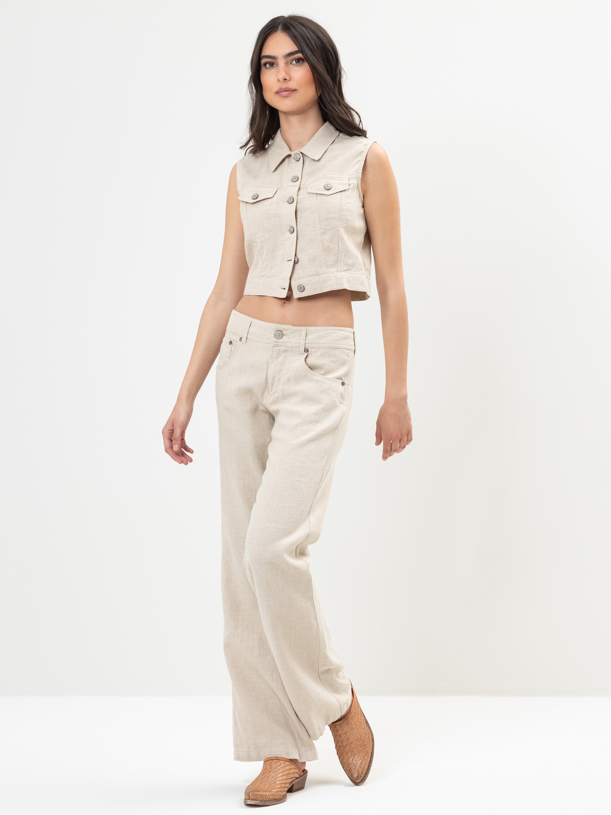 Eine Frau, die eine ärmellose, beigefarbene Weste und eine passende Hose mit hoher Taille und weitem Bein trägt, posiert vor einem schlichten weißen Hintergrund. Sie hat langes dunkles Haar und trägt geflochtene hellbraune Schuhe.