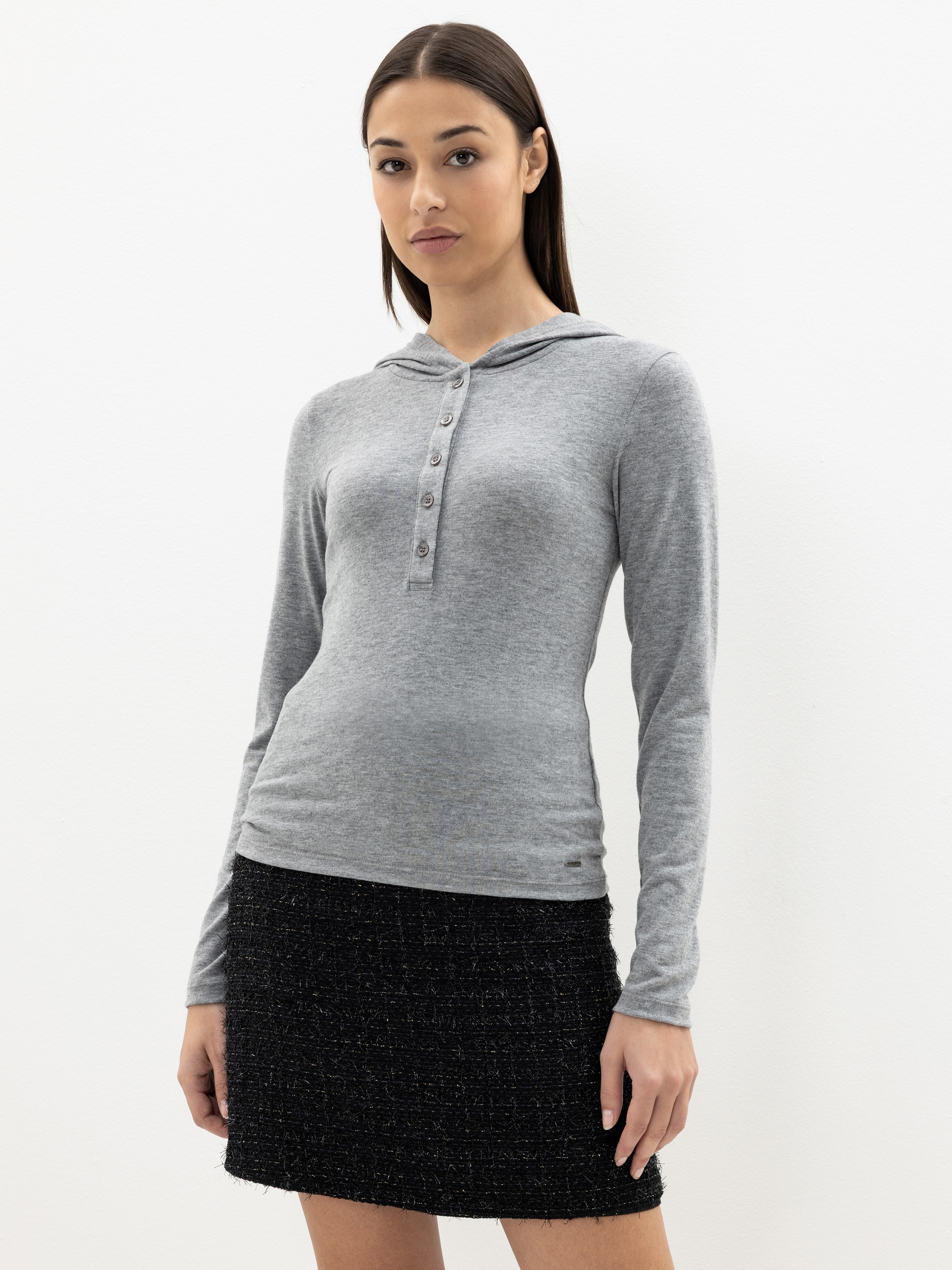 Eine Frau steht vor einem schlichten weißen Hintergrund und trägt ein hellgraues, langärmeliges Kapuzenoberteil mit Knöpfen und einen schwarzen, strukturierten Rock. Sie hat glattes dunkles Haar und einen neutralen Gesichtsausdruck.