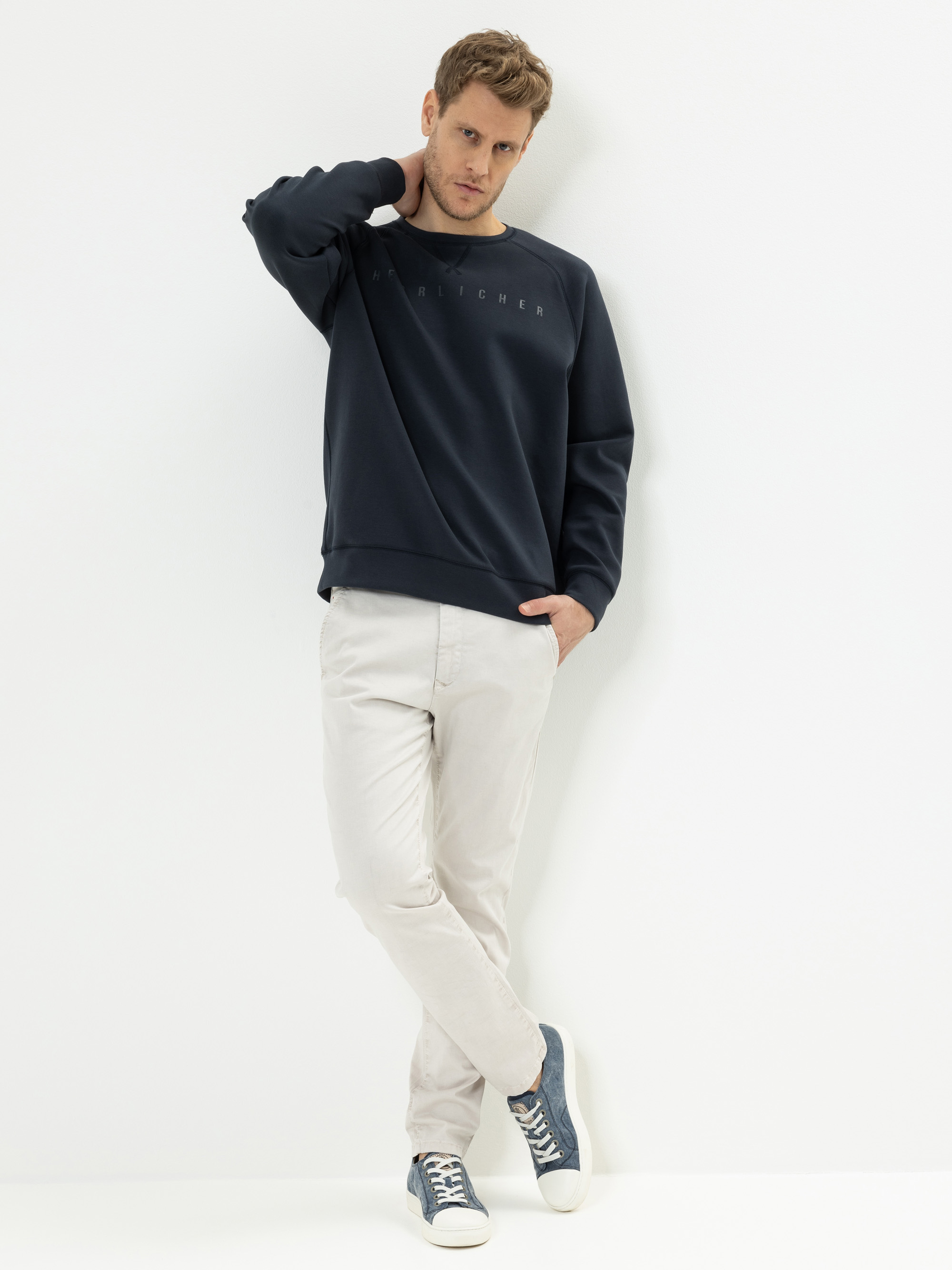 Ein Mann steht vor einer weißen Wand und trägt ein marineblaues Sweatshirt, eine hellbeige Hose und blaue Turnschuhe. Er hat eine Hand hinter dem Kopf und kreuzt seine Beine an den Knöcheln, wobei er leicht nach unten schaut.