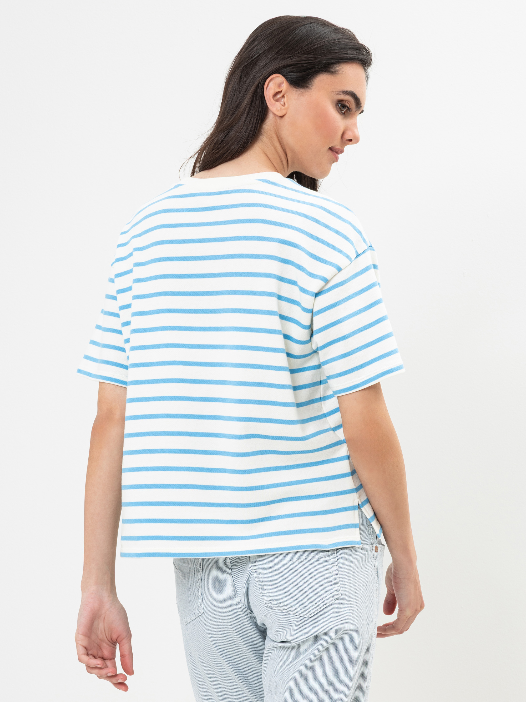 Eine Frau mit langen dunklen Haaren, die ein lockeres weißes T-Shirt mit hellblauen Querstreifen und hellblaue Jeans trägt, wird von hinten gezeigt, wie sie vor einem schlichten weißen Hintergrund über ihre Schulter schaut.