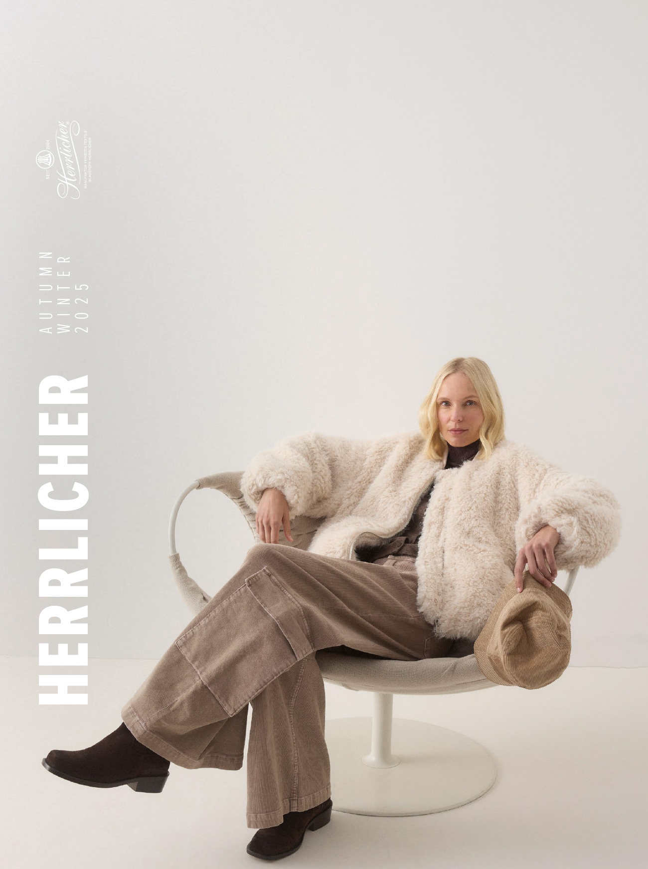 Eine Frau mit blondem Haar sitzt auf einem modernen weißen Stuhl, trägt eine flauschige cremefarbene Jacke, eine braune Hose mit weitem Bein und dunkle Schuhe und hält einen hellbraunen Hut. Der Text auf der linken Seite lautet HERRLICHER HERBST WINTER