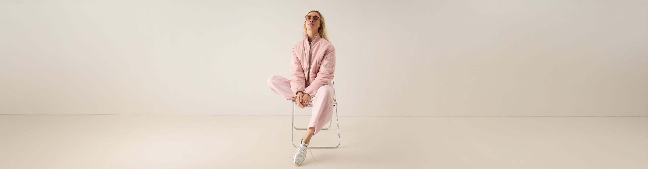 Eine Person, die eine hellrosa Jacke und eine dazu passende Hose trägt, sitzt auf einem einfachen Stuhl in einem minimalistischen, hellen Raum und blickt nach oben, wobei sie die Hände in den Schoß legt und die Beine am Fußgelenk überkreuzt.