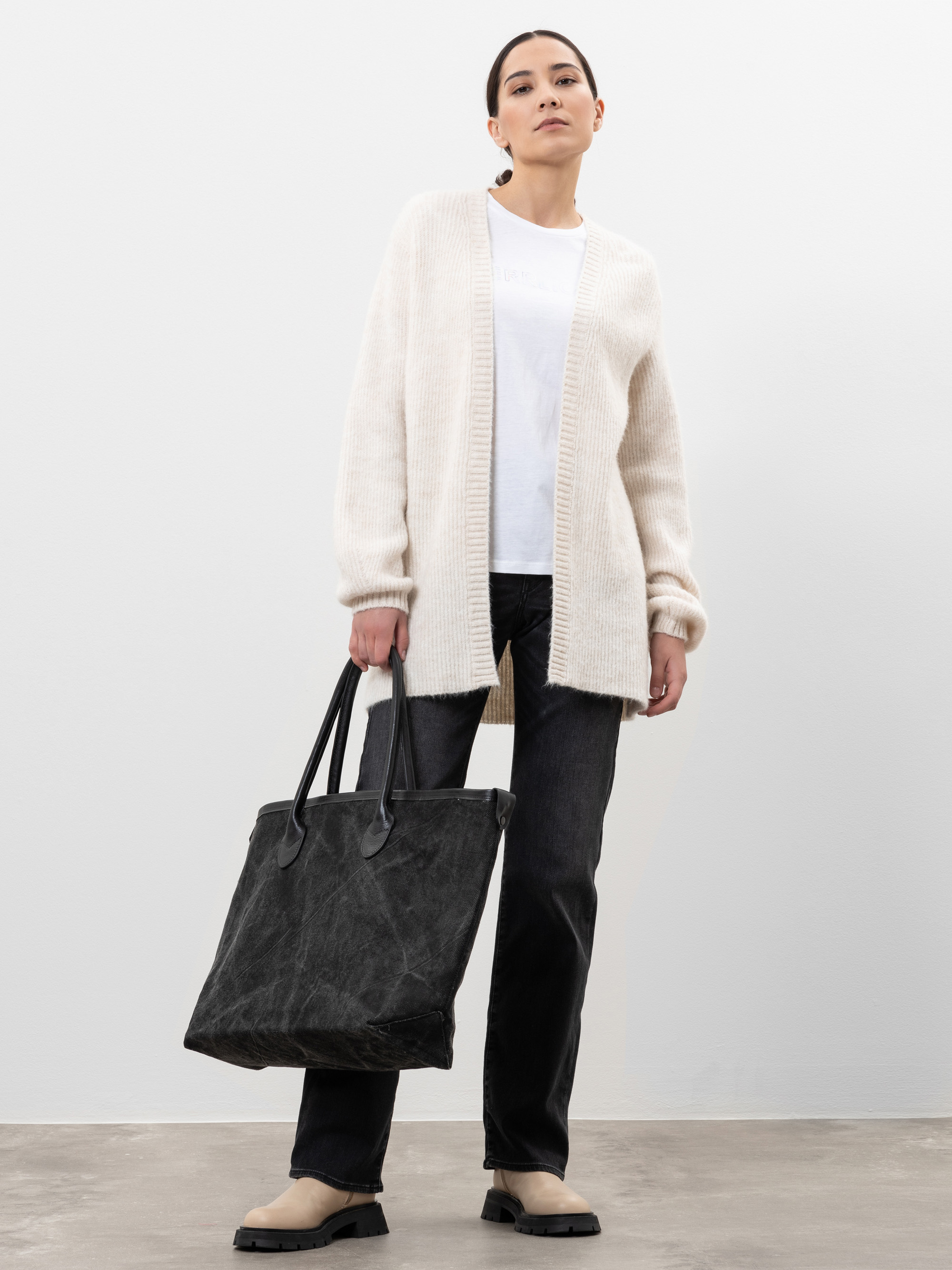 Eine Frau steht vor einem schlichten Hintergrund, trägt eine cremefarbene Strickjacke, ein weißes T-Shirt, schwarze Jeans, beigefarbene Schuhe und hält eine große schwarze Tragetasche in ihrer linken Hand.