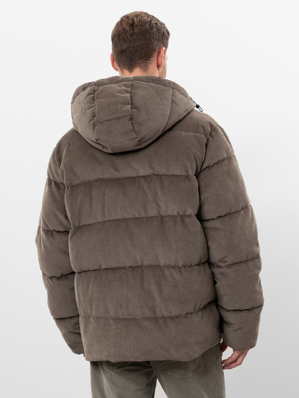 Ein Mann wird von hinten gezeigt, der eine braune Kord-Pulloverjacke mit Kapuze trägt. Er hat kurzes braunes Haar und steht vor einem schlichten weißen Hintergrund.