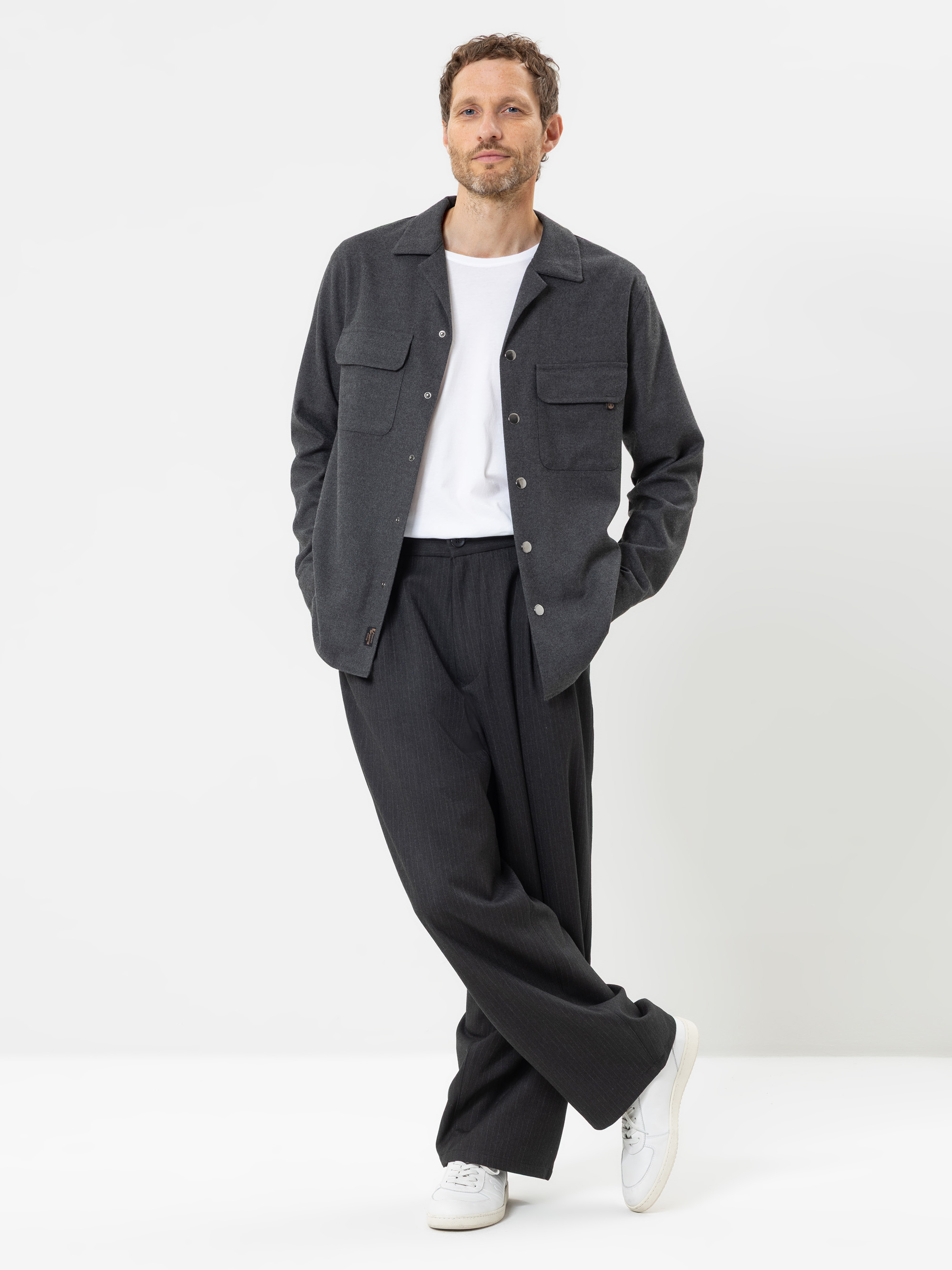 Vor einem weißen Hintergrund steht ein Mann, der eine dunkelgraue Jacke mit Knöpfen, ein weißes T-Shirt, eine weite dunkelgraue Hose und weiße Turnschuhe trägt. Er hat kurzes braunes Haar und einen Bart, eine Hand steckt in der Tasche und er sitzt mi