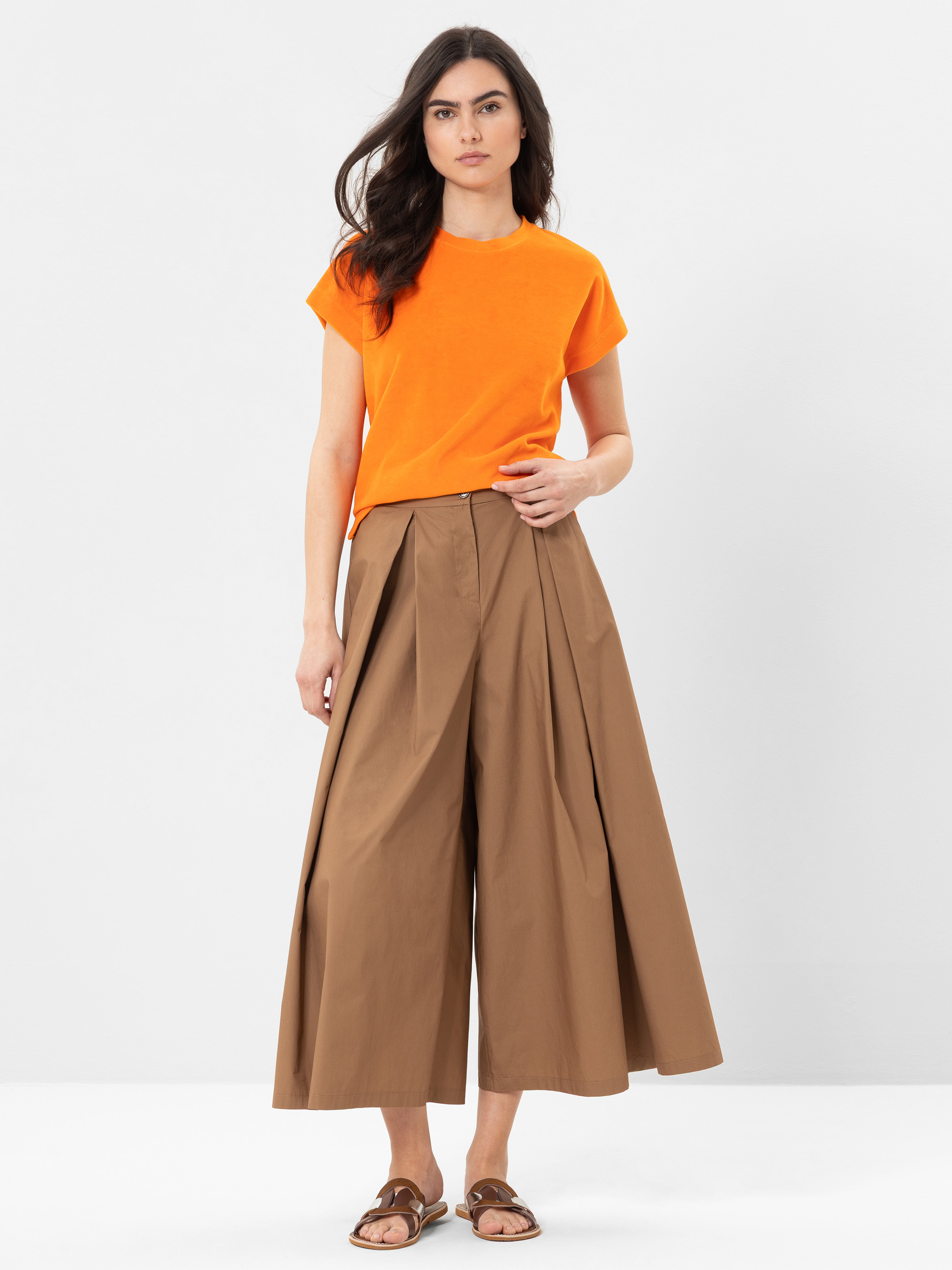 Eine Frau steht vor einem schlichten weißen Hintergrund und trägt ein orangefarbenes kurzärmeliges Oberteil, weite braune Plissee-Culottes und braune Sandalen. Sie hat langes dunkles Haar und einen neutralen Gesichtsausdruck.