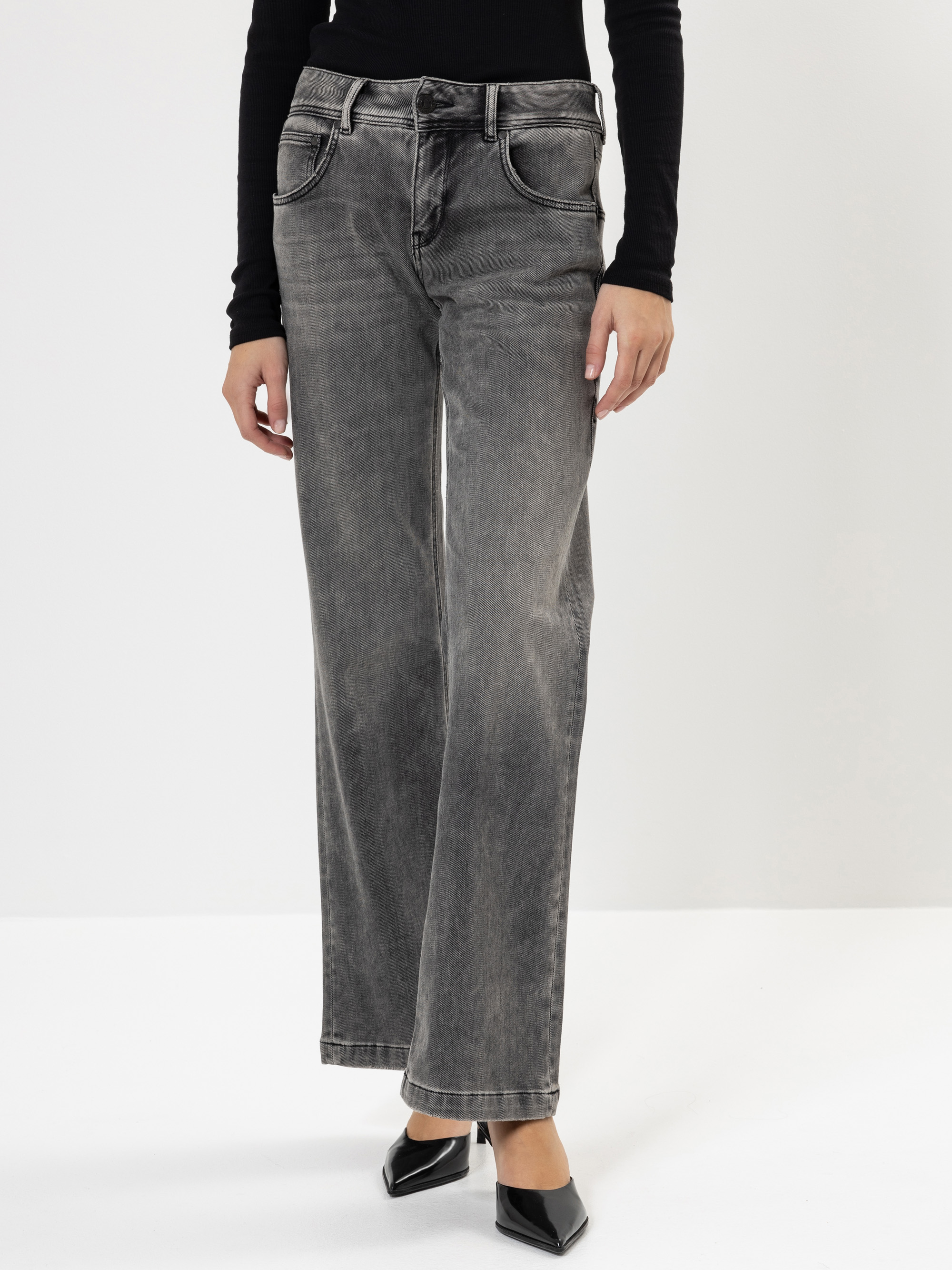 Eine Person, die eine graue Jeans mit hoher Taille und weitem Bein, ein schwarzes, langärmeliges Oberteil und schwarze, spitze Schuhe trägt, steht vor einem schlichten weißen Hintergrund.