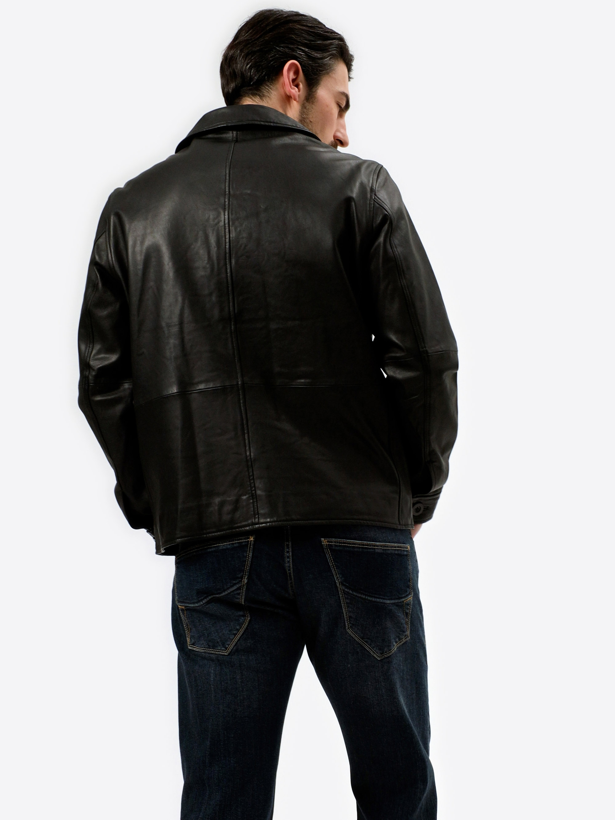 Ein Mann mit dunklem Haar ist von hinten zu sehen, er trägt eine schwarze Lederjacke und dunkelblaue Jeans vor einem schlichten weißen Hintergrund.