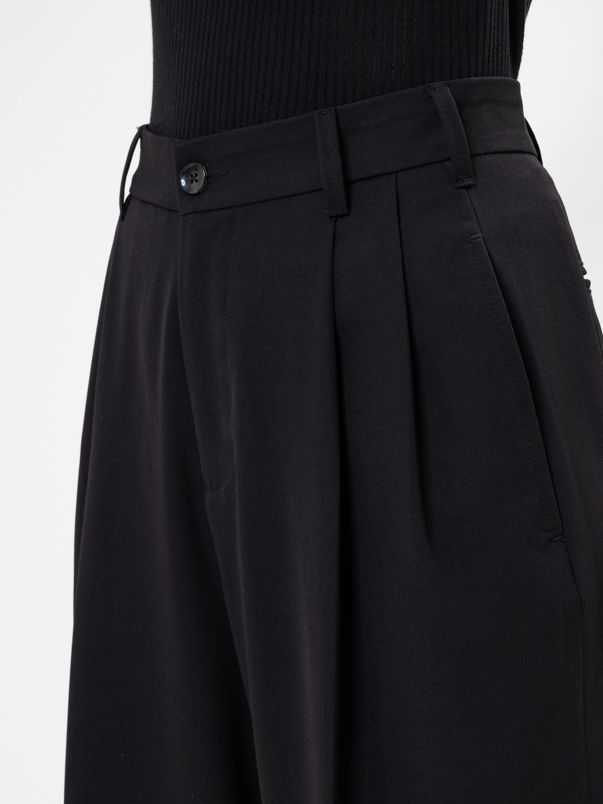 Nahaufnahme einer Person, die eine schwarze Bundfaltenhose mit hoher Taille, Gürtelschlaufen und Knopfverschluss trägt, dazu ein schwarzes geripptes Oberteil, das in die Hose gesteckt wird. Der Hintergrund ist einfarbig weiß.