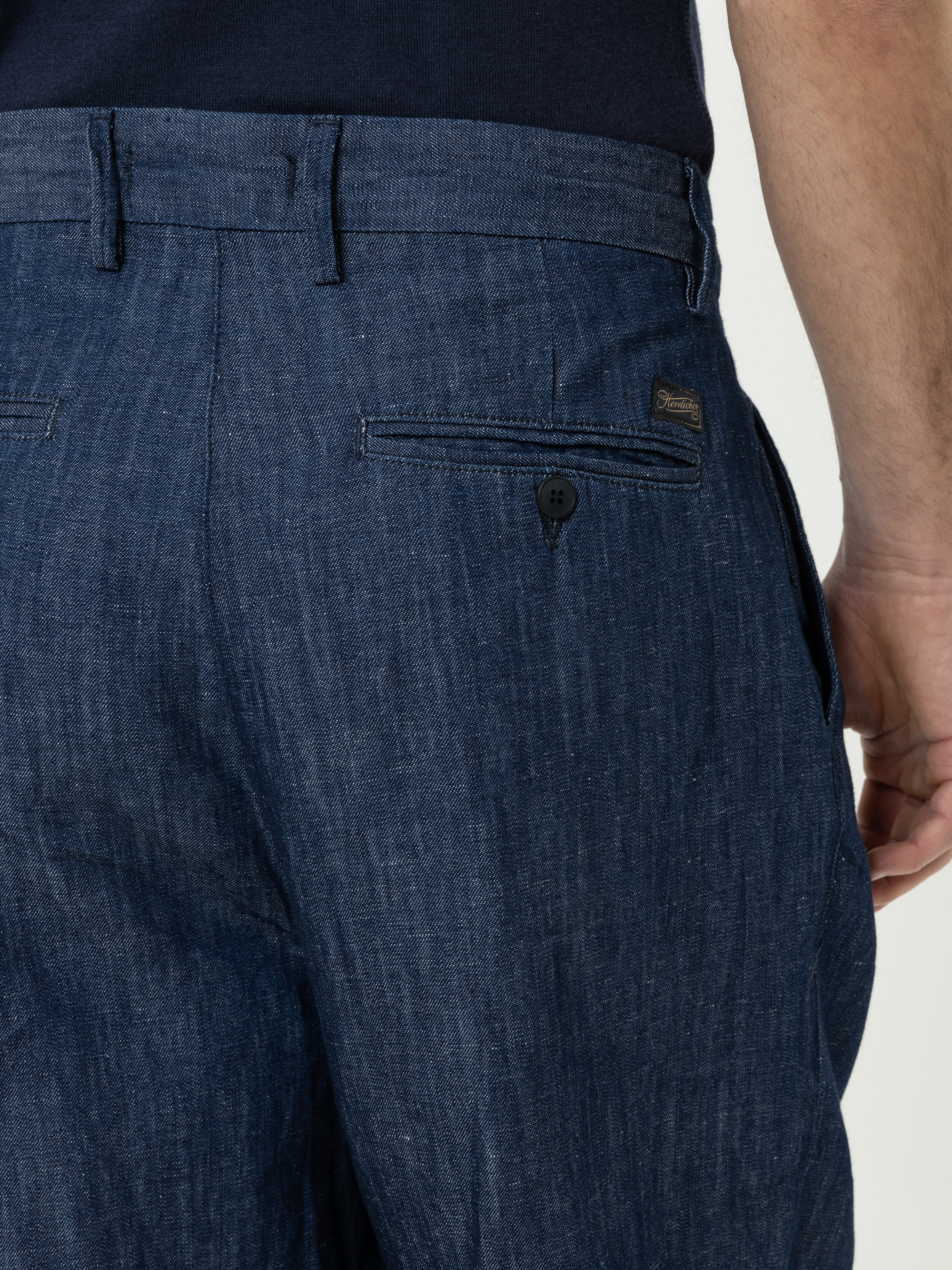 Nahaufnahme einer Person, die eine dunkelblaue Jeanshose mit einer durch einen Knopf gesicherten Gesäßtasche trägt. Der Arm der Person ist am rechten Rand des Bildes zu sehen.