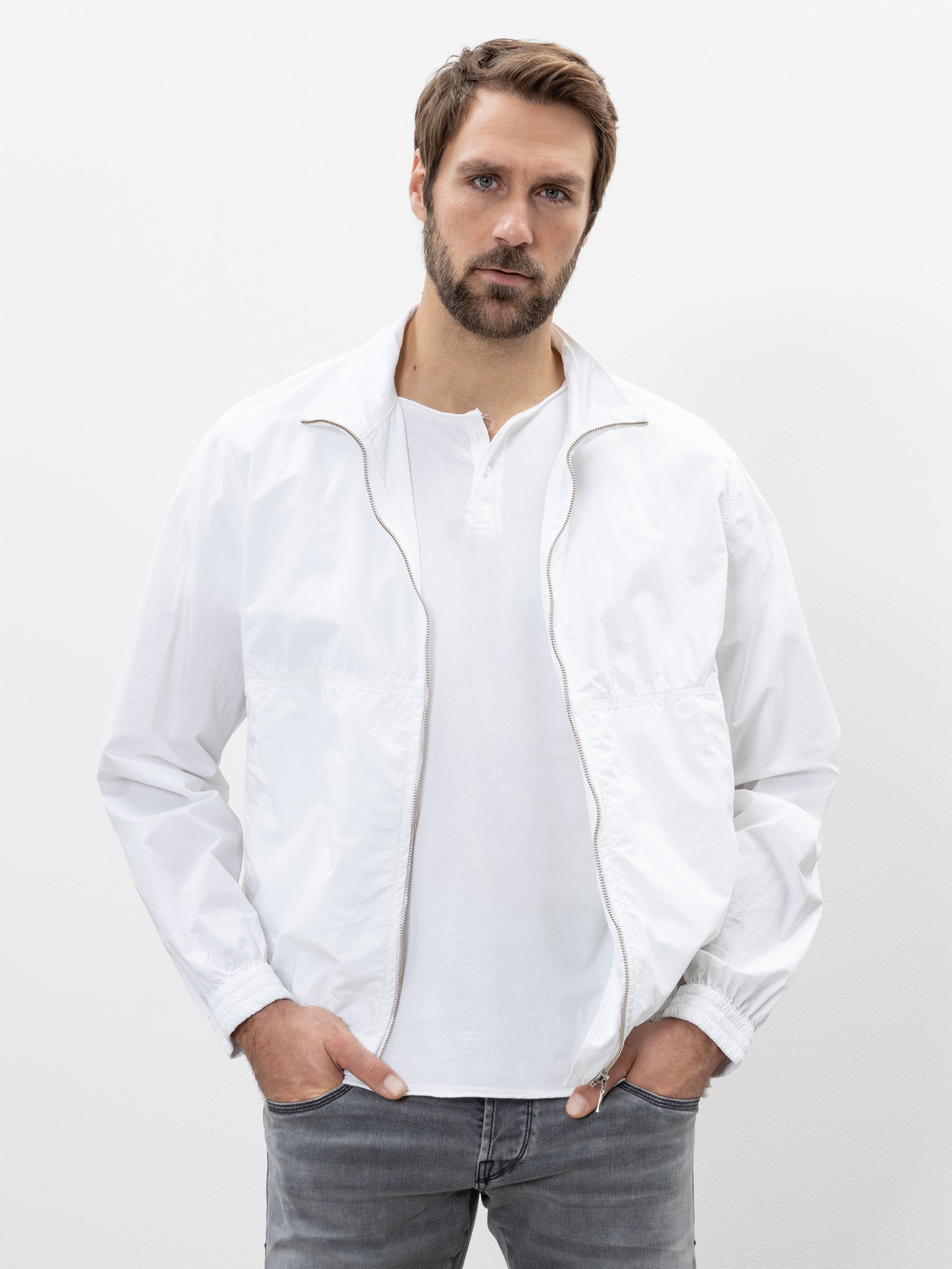 Ein Mann mit kurzen braunen Haaren und Bart trägt eine weiße Jacke mit Reißverschluss über einem weißen Hemd und graue Jeans. Er steht vor einem schlichten weißen Hintergrund und hat die Hände in den Taschen.