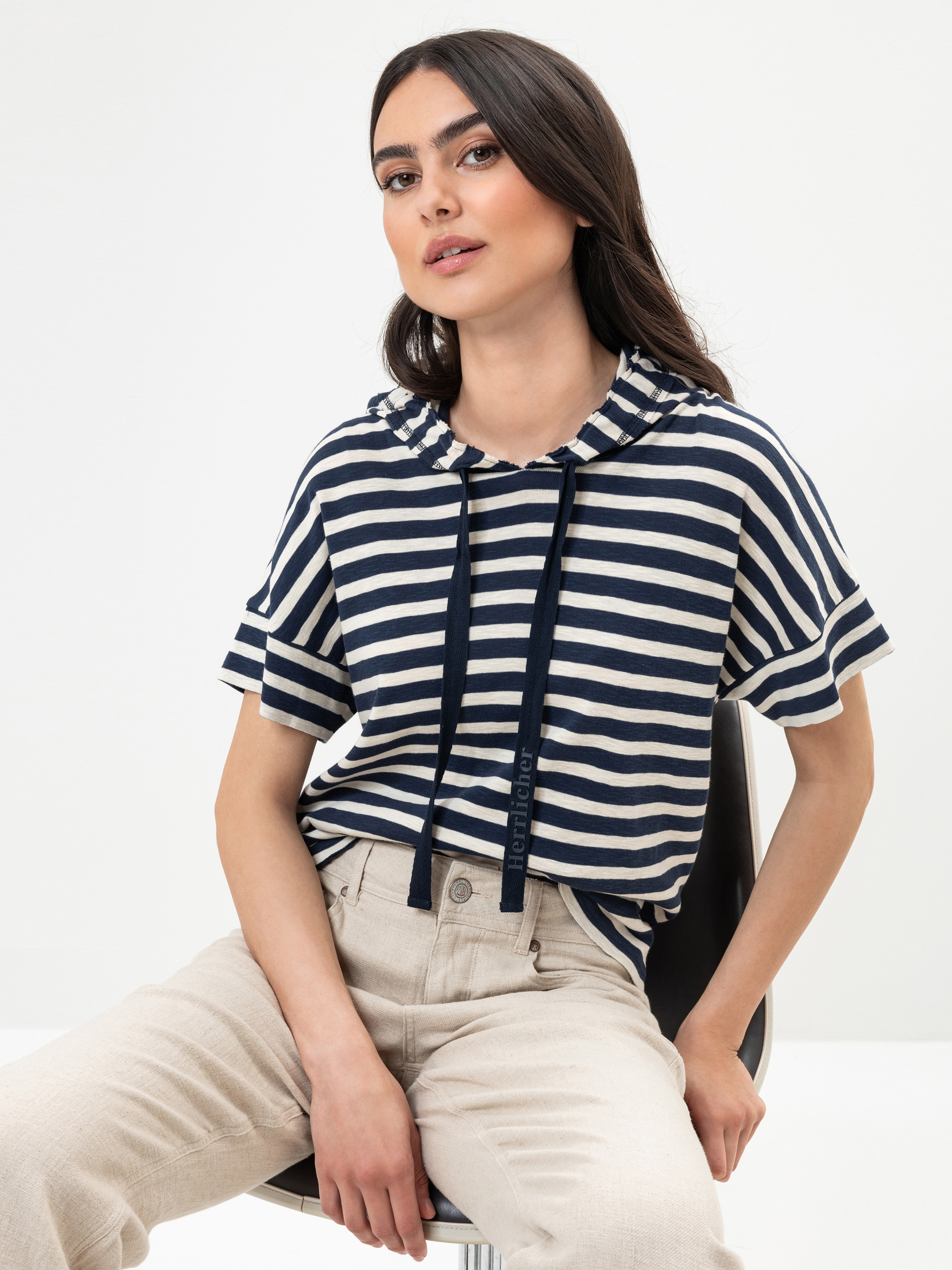 Eine Frau mit langen dunklen Haaren sitzt auf einem Stuhl, trägt einen marineblau-weiß gestreiften kurzärmeligen Kapuzenpulli und eine hellbeige Hose und posiert vor einem schlichten weißen Hintergrund.