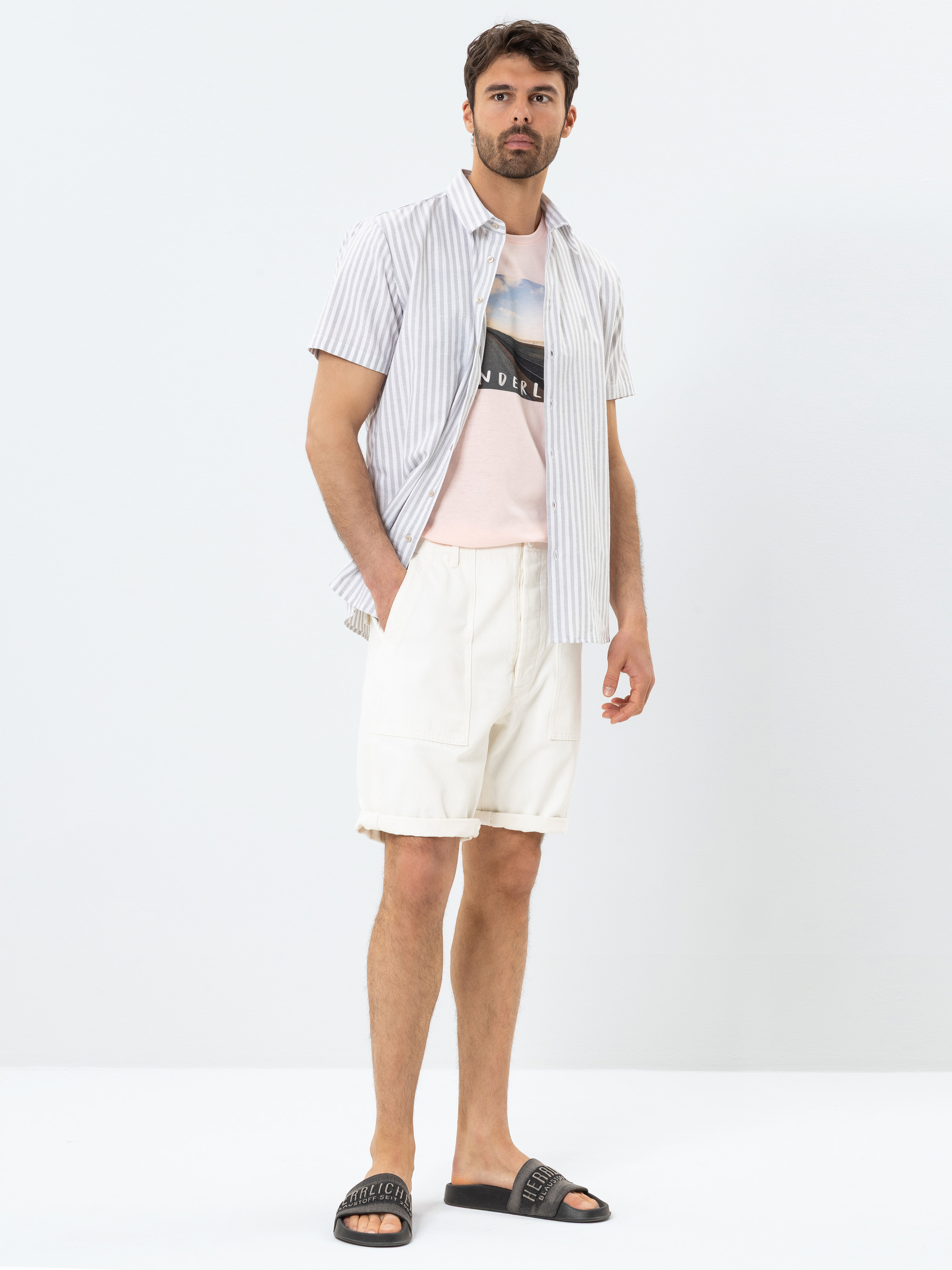 Ein Mann steht vor einem schlichten weißen Hintergrund und trägt ein kurzärmeliges gestreiftes Button-up-Hemd über einem grafischen T-Shirt, weiße Shorts und schwarze Slide-Sandalen. Er hat dunkles Haar und einen gestutzten Bart und blickt leicht zur