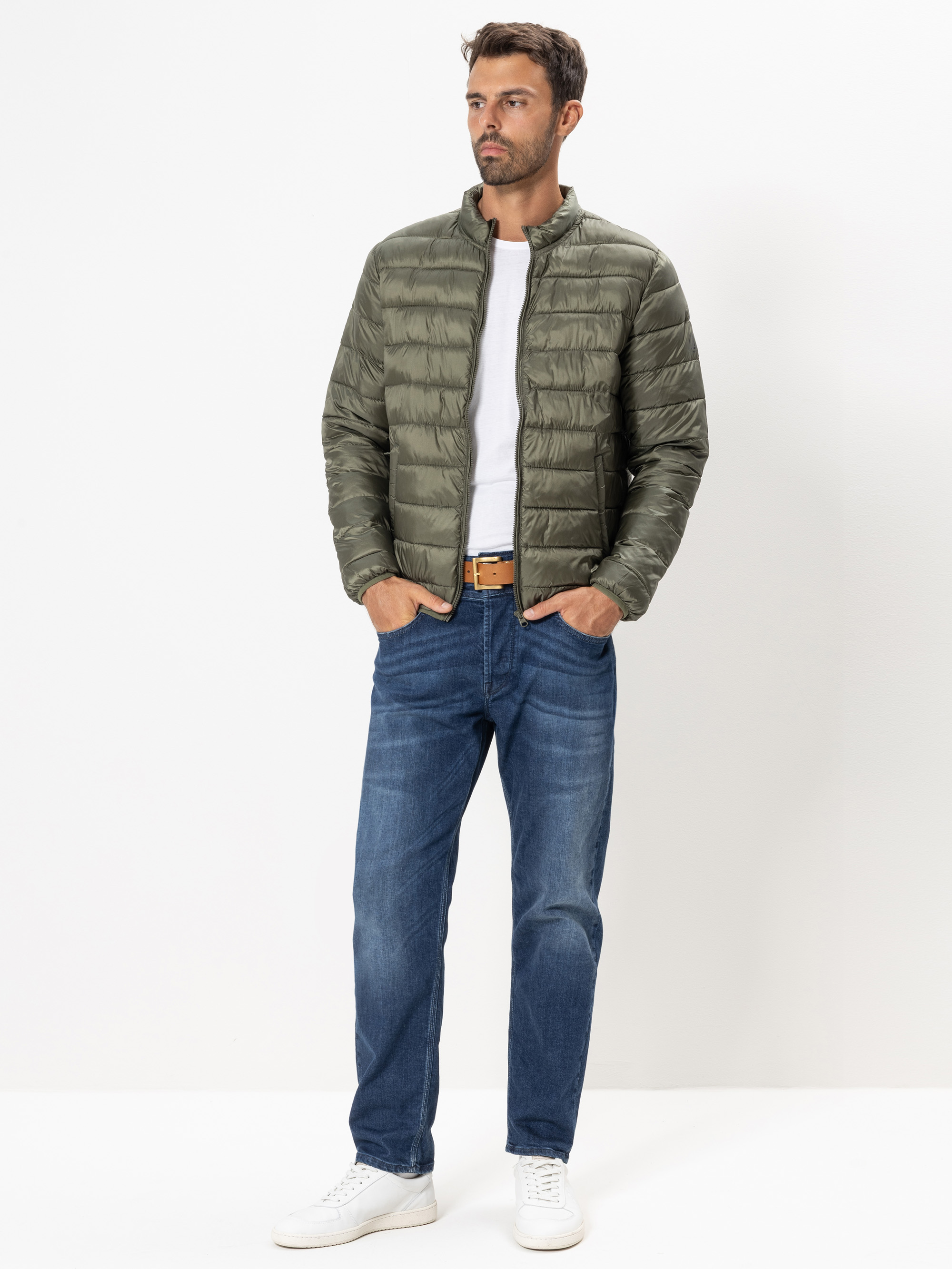 Ein Mann steht vor einem schlichten weißen Hintergrund und trägt eine grüne Pufferjacke, ein weißes T-Shirt, blaue Jeans, einen hellbraunen Gürtel und weiße Turnschuhe. Seine Hände stecken in den Jackentaschen und er schaut zur Seite.