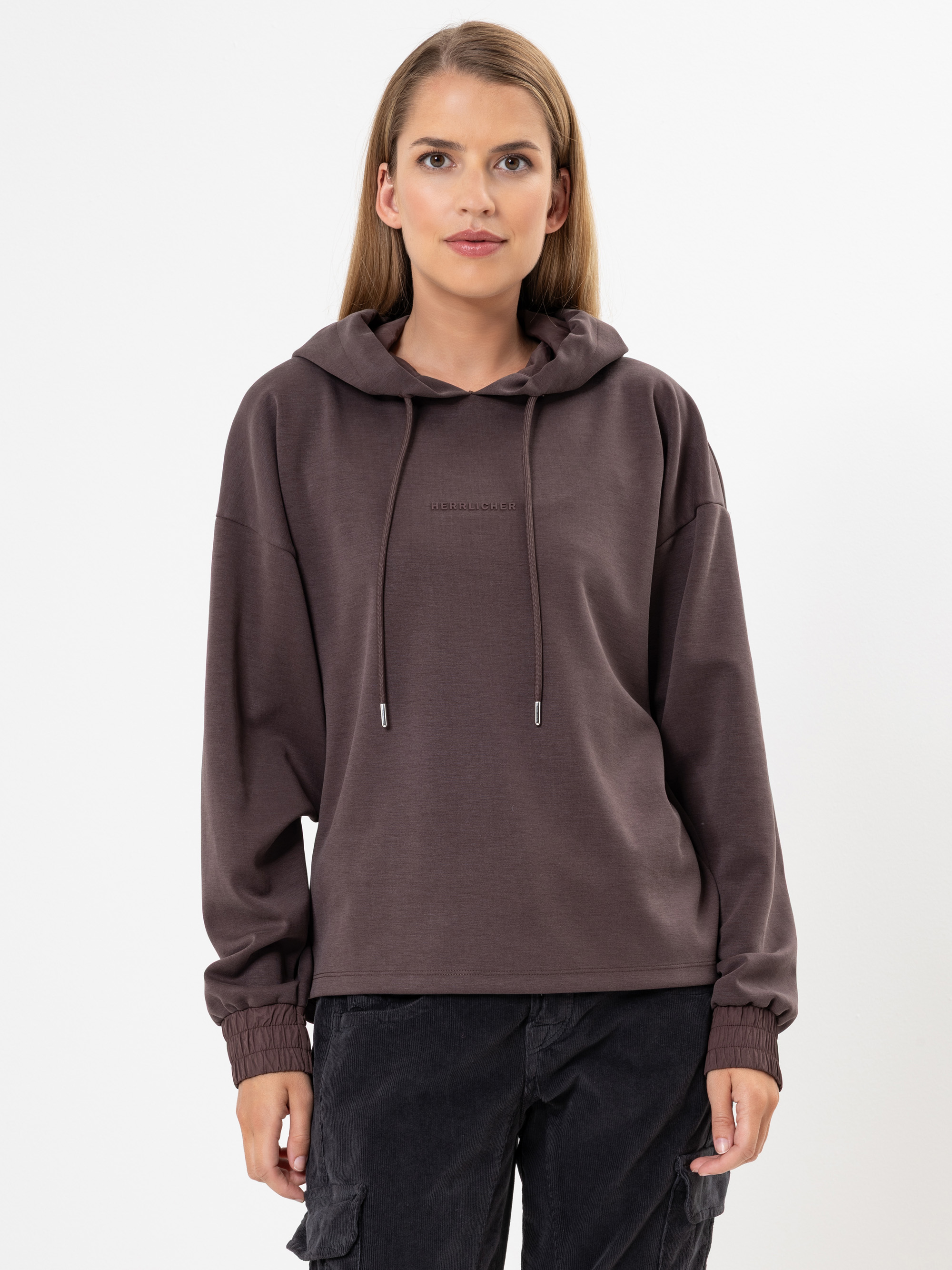 Eine Frau mit langen hellbraunen Haaren trägt einen weiten, dunkelbraunen Kapuzenpulli mit Kordelzug und eine lässige schwarze Hose. Sie steht vor einem schlichten weißen Hintergrund.
