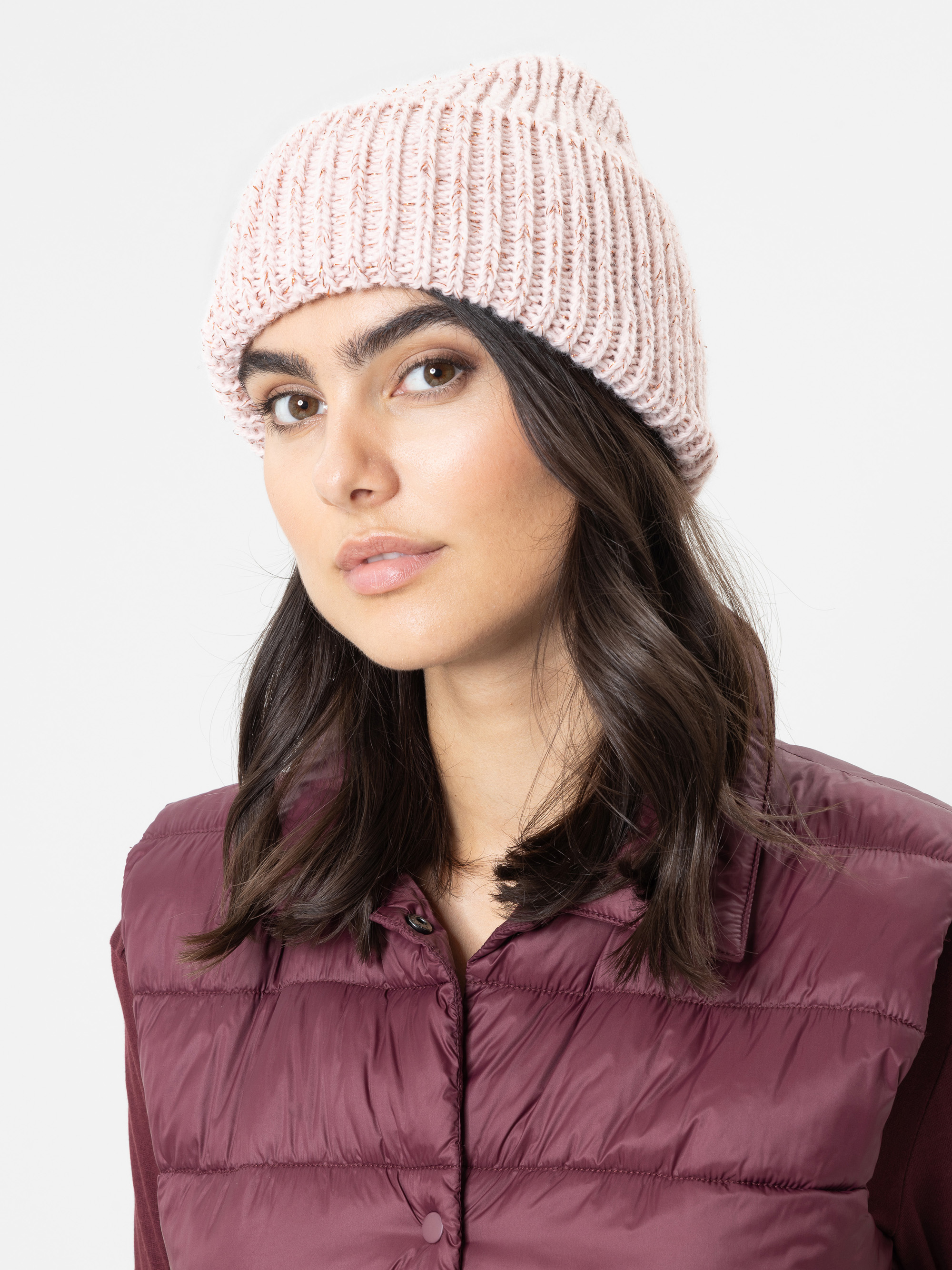 Eine Frau mit mittellangem dunkelbraunem Haar, die eine hellrosa Strickmütze und eine burgunderrote Daunenweste über einem farblich passenden Shirt trägt und mit neutralem Gesichtsausdruck vor einem schlichten hellen Hintergrund in die Kamera blickt.