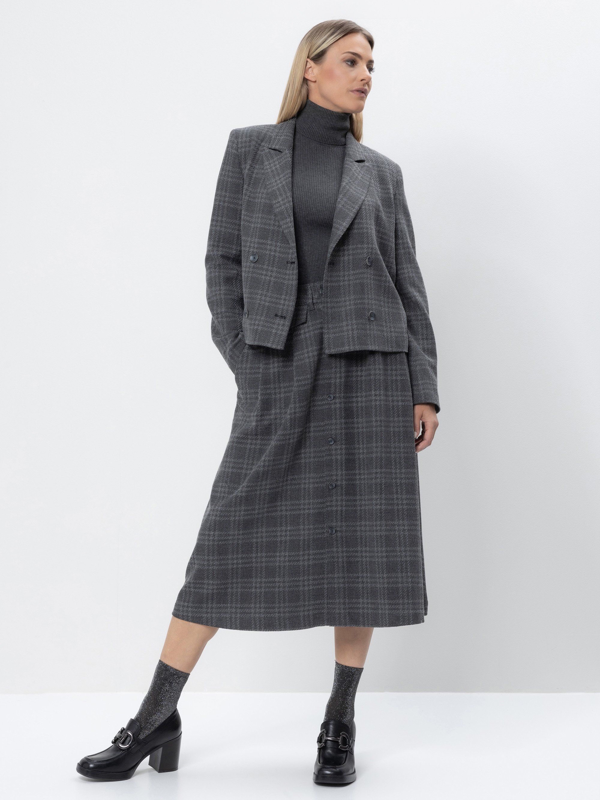 Eine Frau steht vor einem weißen Hintergrund und trägt einen grau karierten Blazer und einen dazu passenden Midirock, gepaart mit einem dunklen Rollkragenpullover, grauen Socken und schwarzen Halbschuhen mit Absatz. Sie hat glattes blondes Haar und eine