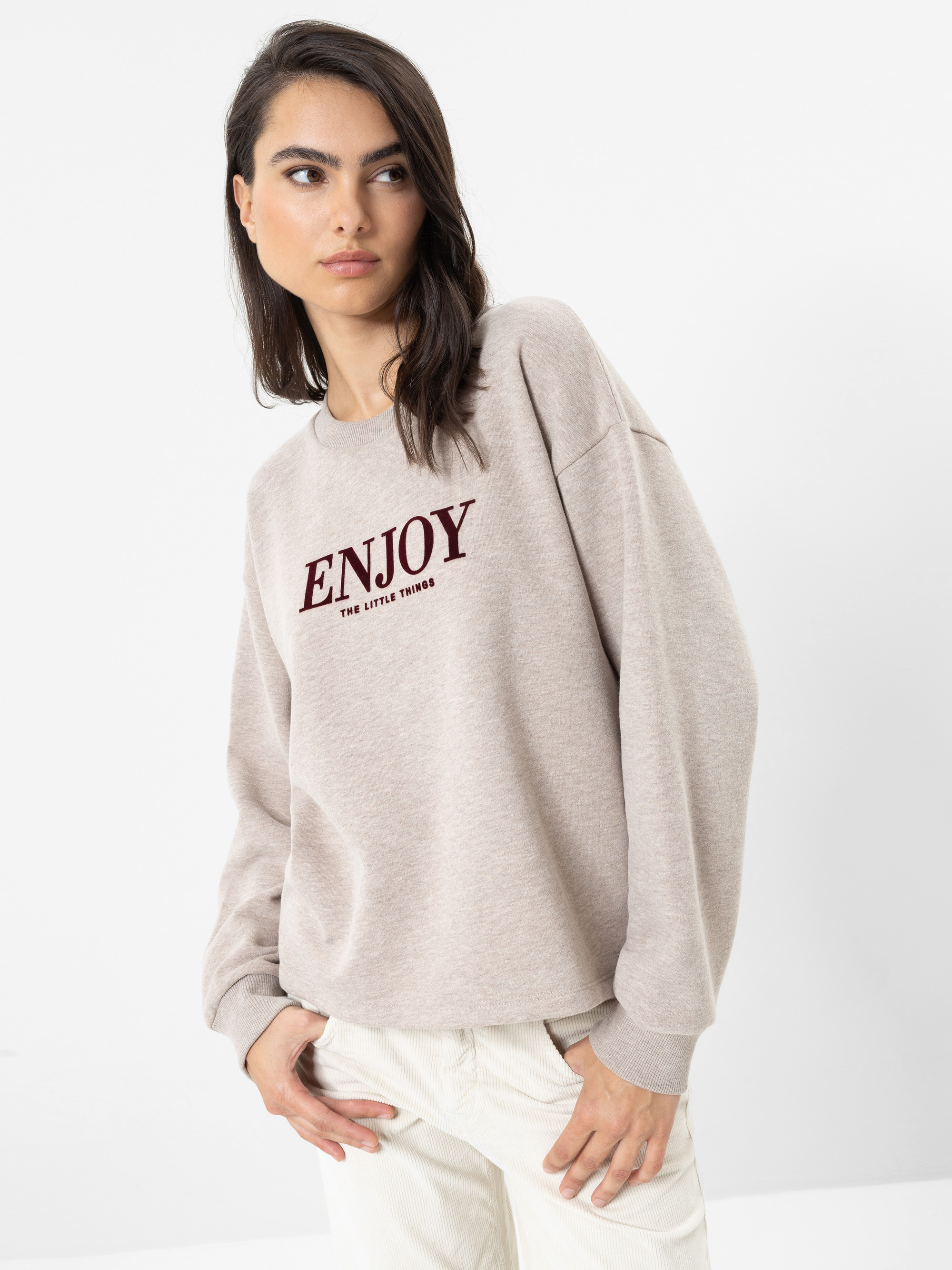 Eine Frau mit langen dunklen Haaren, die einen beigen Pullover mit der Aufschrift „ENJOY the little things“ in roten Buchstaben trägt und dazu eine helle Hose, steht vor einem schlichten weißen Hintergrund und blickt nach links.