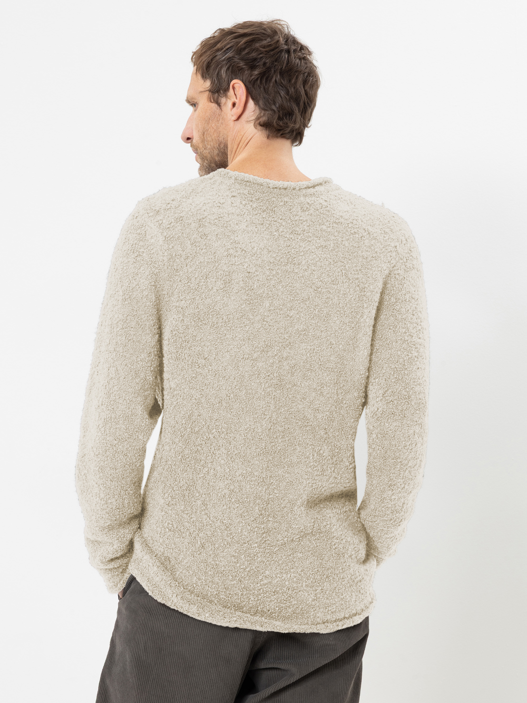Ein Mann mit braunen Haaren, von hinten gesehen, der einen beigen, strukturierten, langärmeligen Strickpullover und eine dunkelgraue Hose trägt, vor einem schlichten weißen Hintergrund.