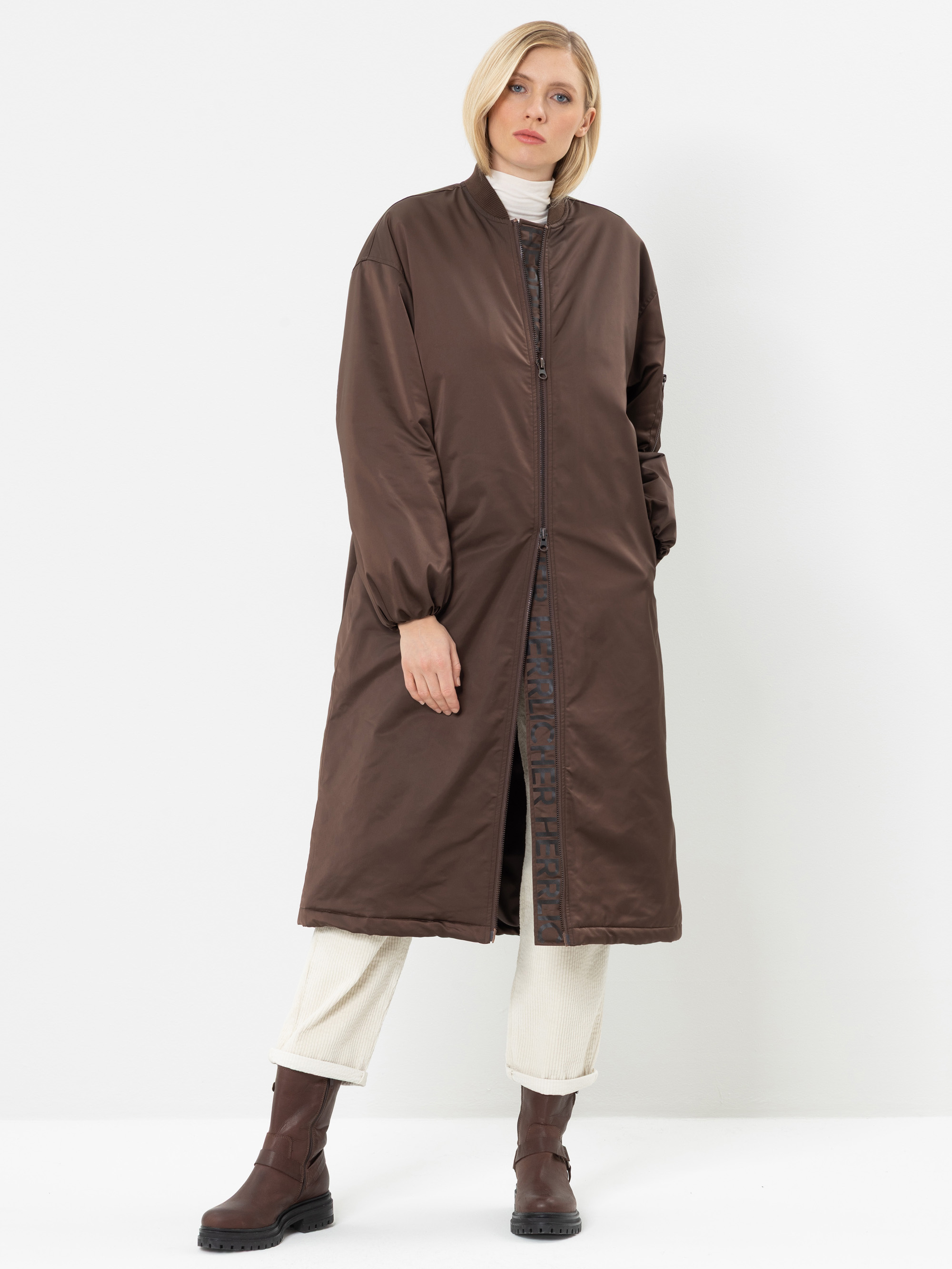 Eine Person, die einen langen, braunen Steppmantel über einem weißen Rollkragenpullover und einer cremefarbenen Hose trägt und dazu braune Stiefel anhat, steht vor einem schlichten weißen Hintergrund.
