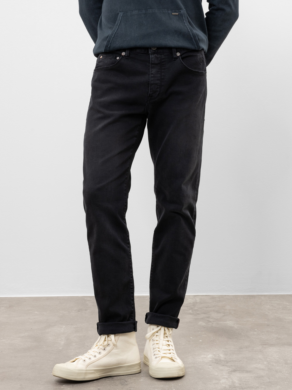 Eine Person, die einen dunklen Kapuzenpullover, schwarze Jeans und weiße High-Top-Turnschuhe trägt, steht vor einer schlichten, hellen Wand und einem Betonboden. Nur die untere Hälfte ihres Körpers ist sichtbar.