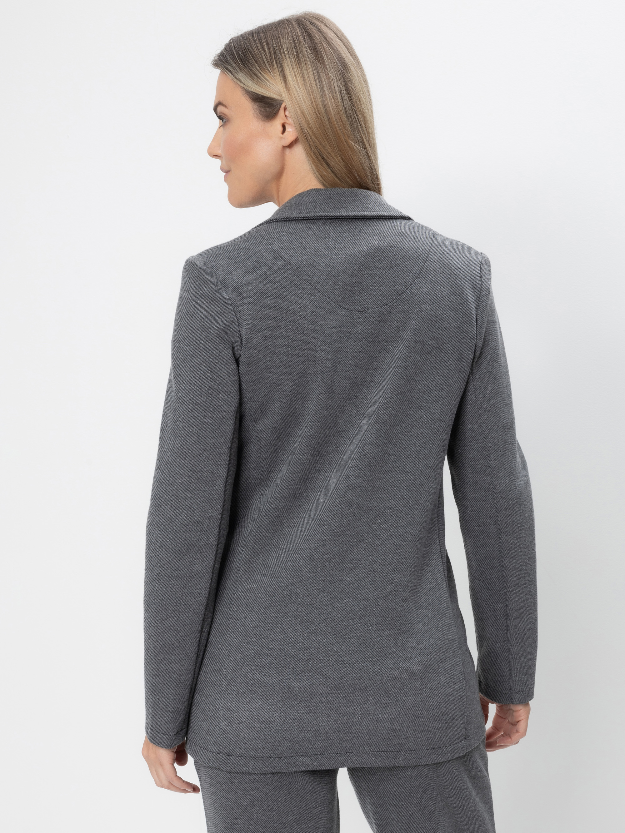 Eine Frau mit glattem, blondem Haar steht mit dem Gesicht zur Wand und trägt einen grauen Blazer und eine passende Hose vor einem schlichten weißen Hintergrund.