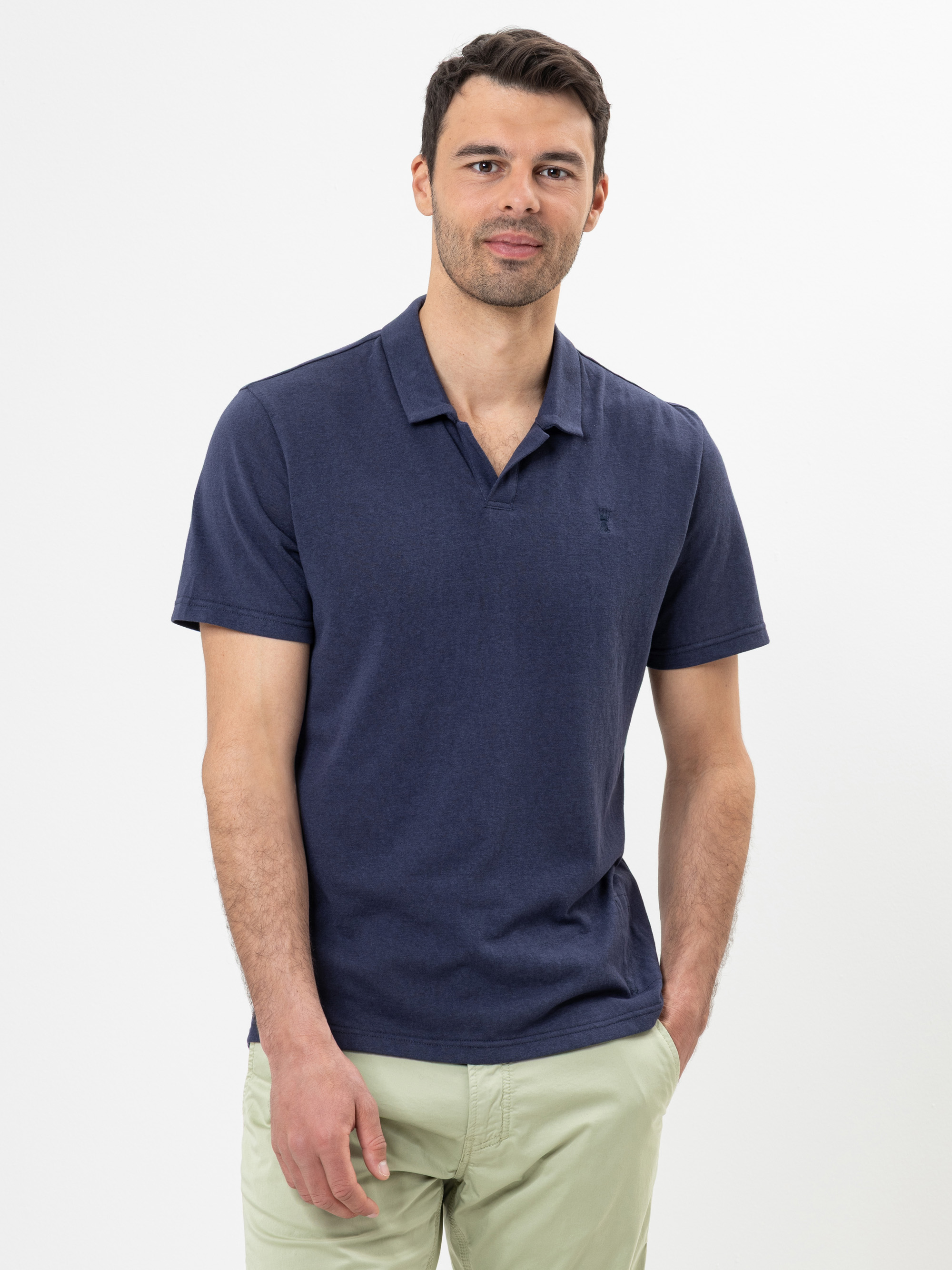 Ein Mann mit kurzen dunklen Haaren und gestutztem Bart trägt ein marineblaues Polohemd und eine hellbeige Hose, steht vor einem schlichten weißen Hintergrund und blickt mit einem leichten Lächeln in die Kamera.