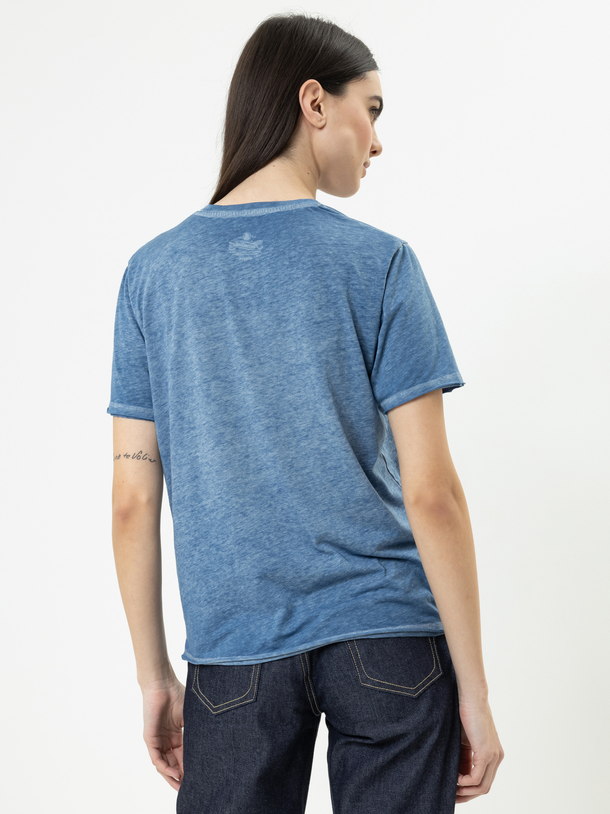 Frau mit langen dunklen Haaren, die ein blaues T-Shirt und dunkle Jeans trägt und mit dem Rücken zur Kamera vor einem schlichten weißen Hintergrund steht. Auf ihrem linken Unterarm ist eine kleine Tätowierung zu sehen.
