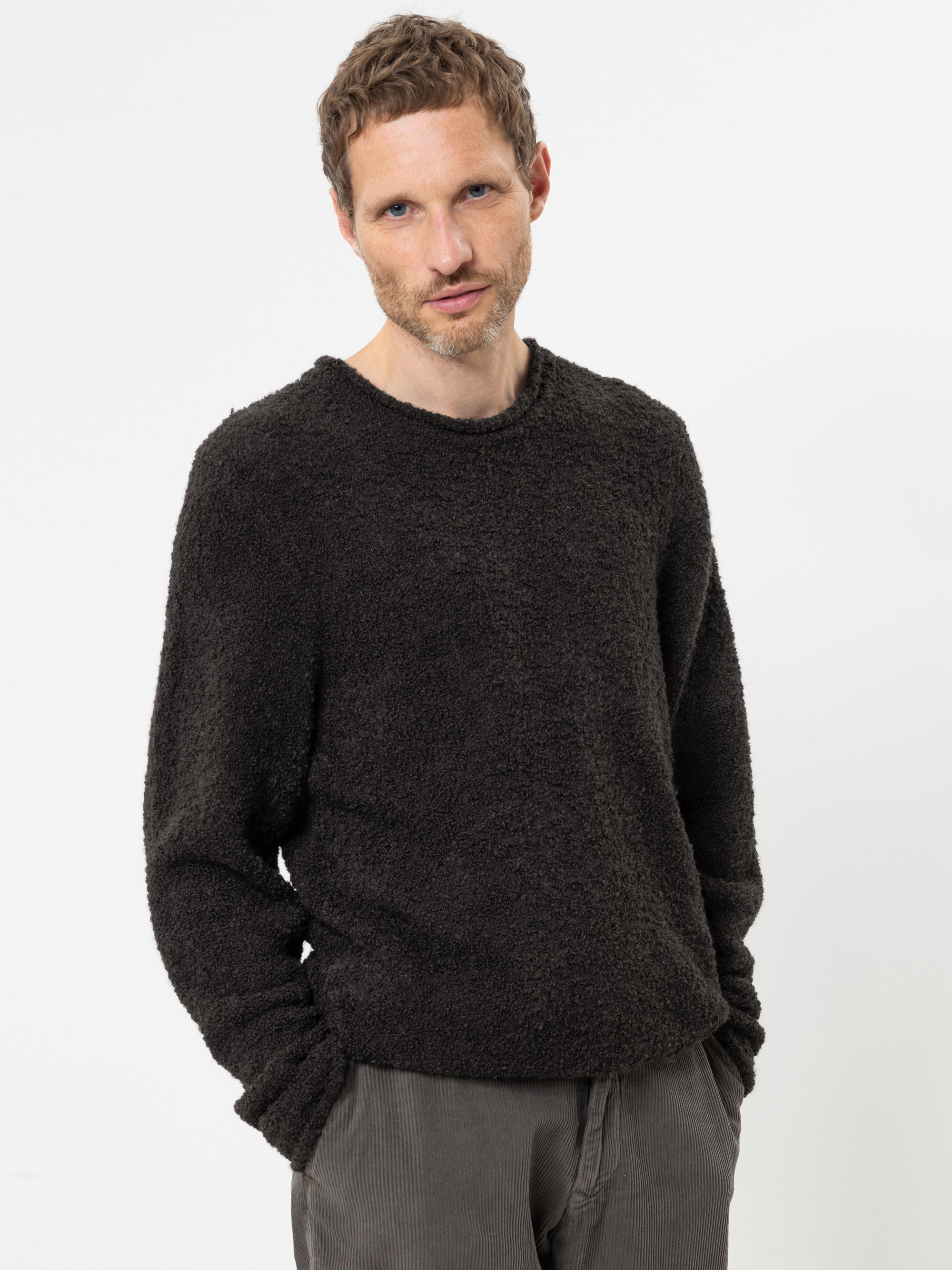 Ein Mann mit kurzen, gewellten Haaren und leichtem Bartstoppeln trägt einen strukturierten, dunklen Pullover und eine graue Hose. Er steht vor einem schlichten weißen Hintergrund, die Hände in den Taschen, und blickt leicht in Richtung Kamera.