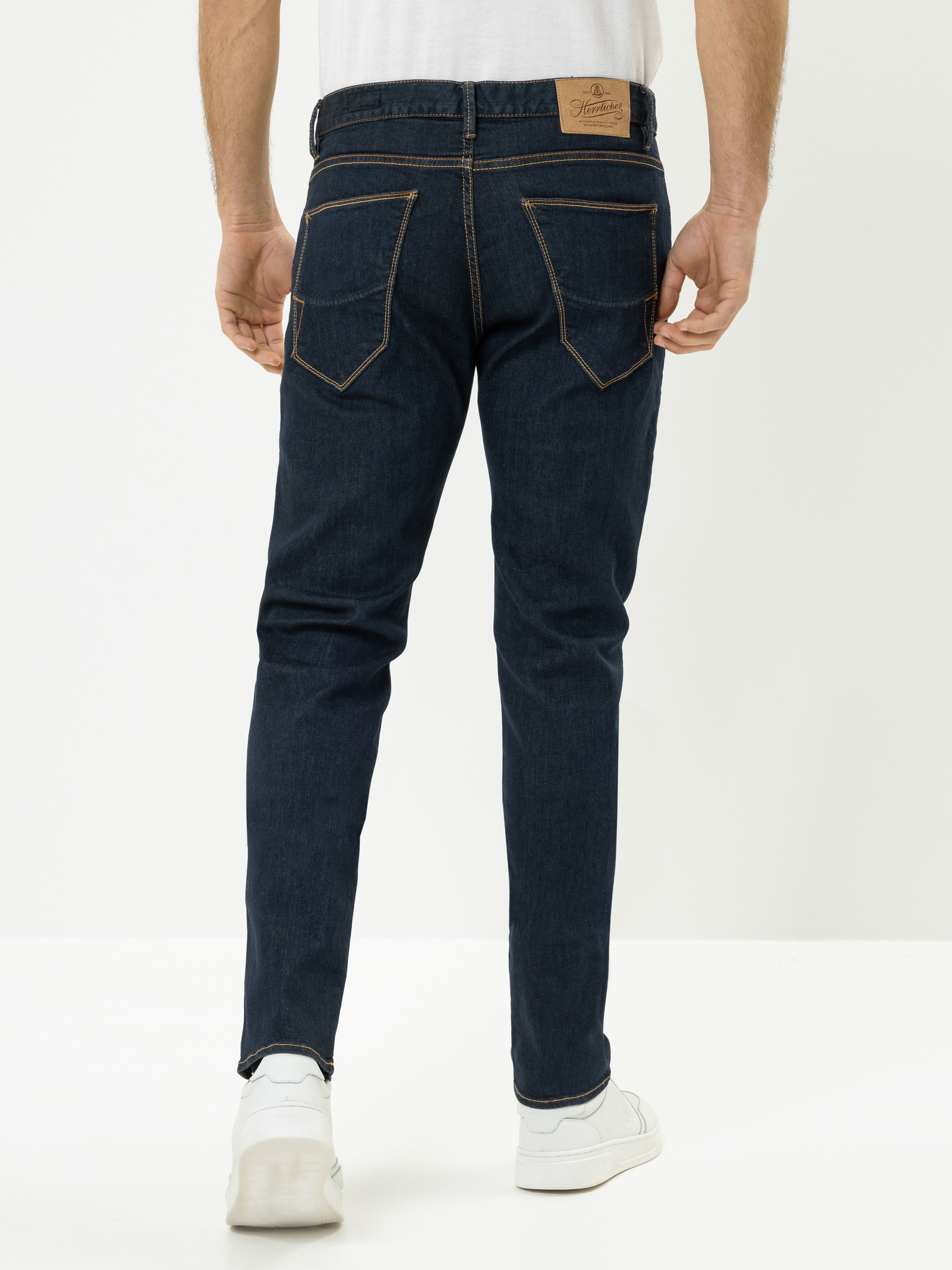 Ein Mann, der ein weißes T-Shirt, eine dunkelblaue Jeans mit braunen Nähten und weiße Turnschuhe trägt, steht mit dem Gesicht nach hinten und zeigt die Rückenansicht der Jeans vor einem schlichten weißen Hintergrund.